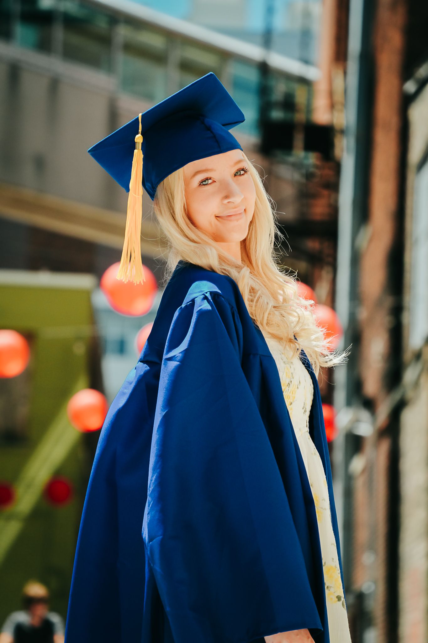 Smiling blonde graduate in blue cap and gown posing outdoors with red lanterns in background.