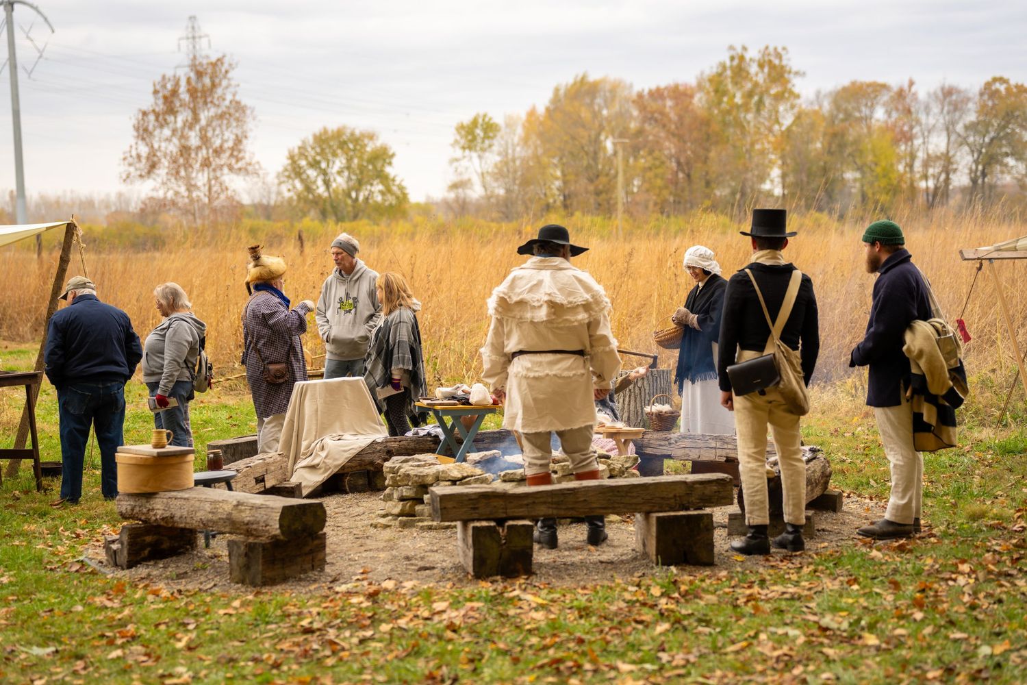 Historical reenactors in 19th century period costumes gather around a campfire in an autumn field setting.