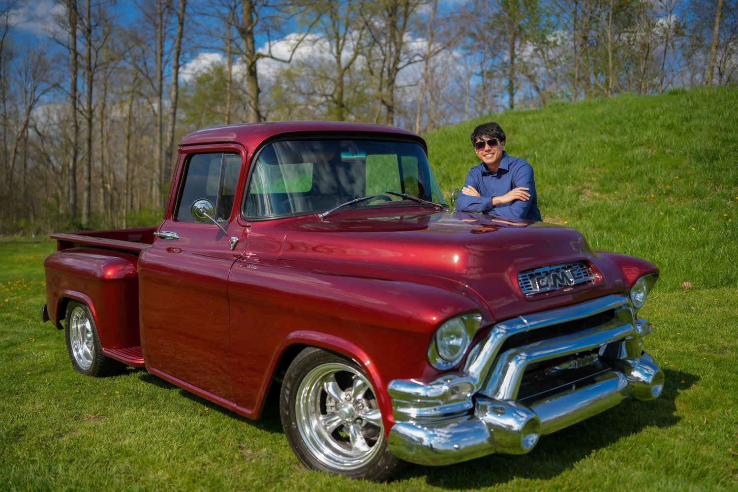 Smiling young man leaning on a classic restored red GMC pickup truck on a green grassy field.