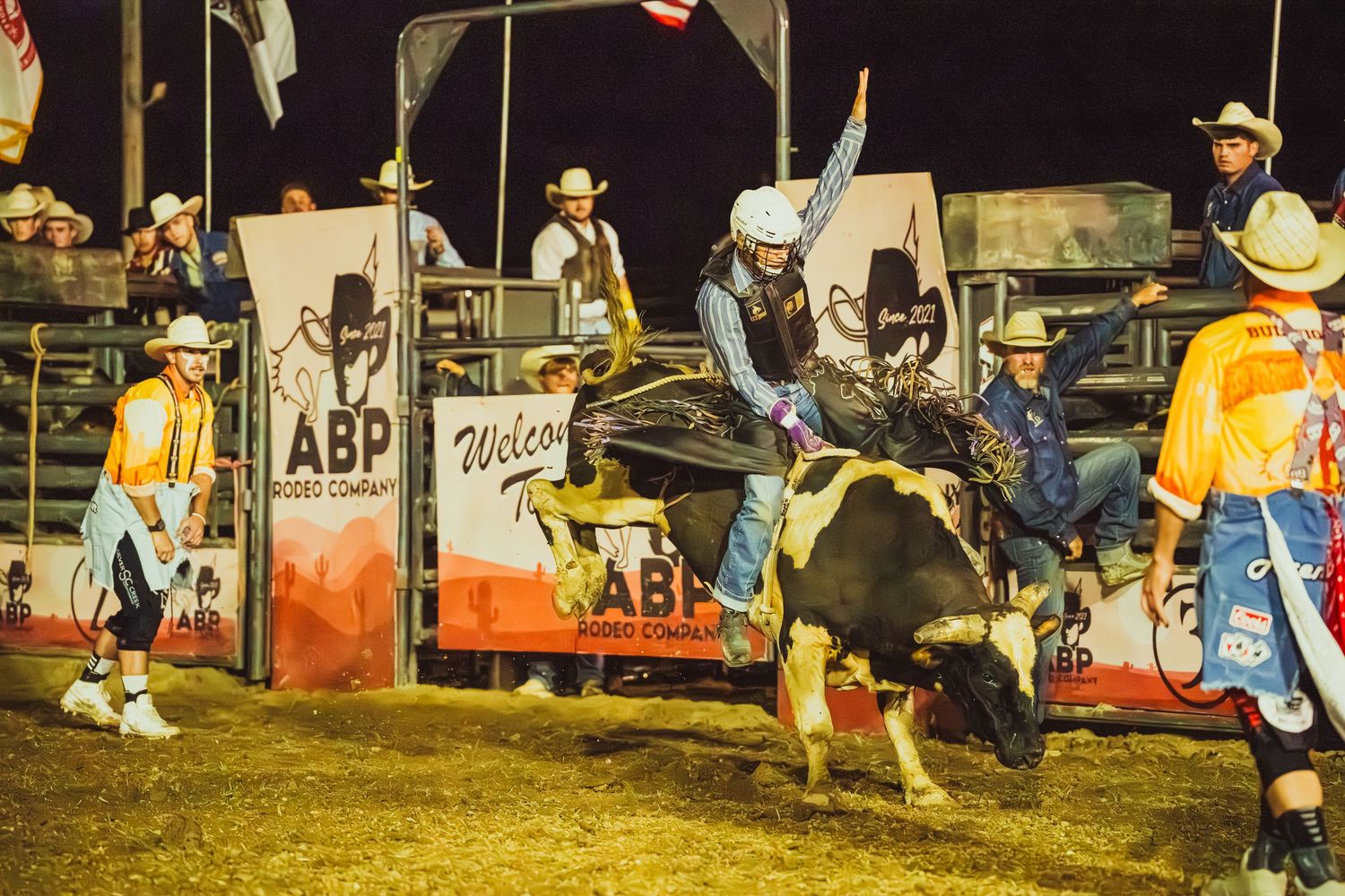 Bull rider competing at ABP Rodeo Cowboys event at night, with rodeo clowns and crowd in background.