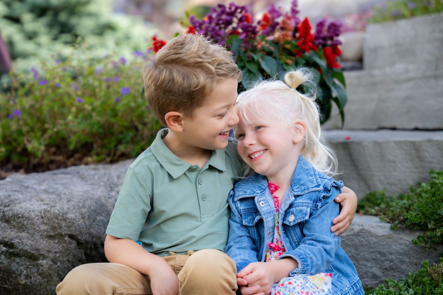Two young siblings smiling and hugging outdoors, surrounded by colorful garden flowers on a sunny day.