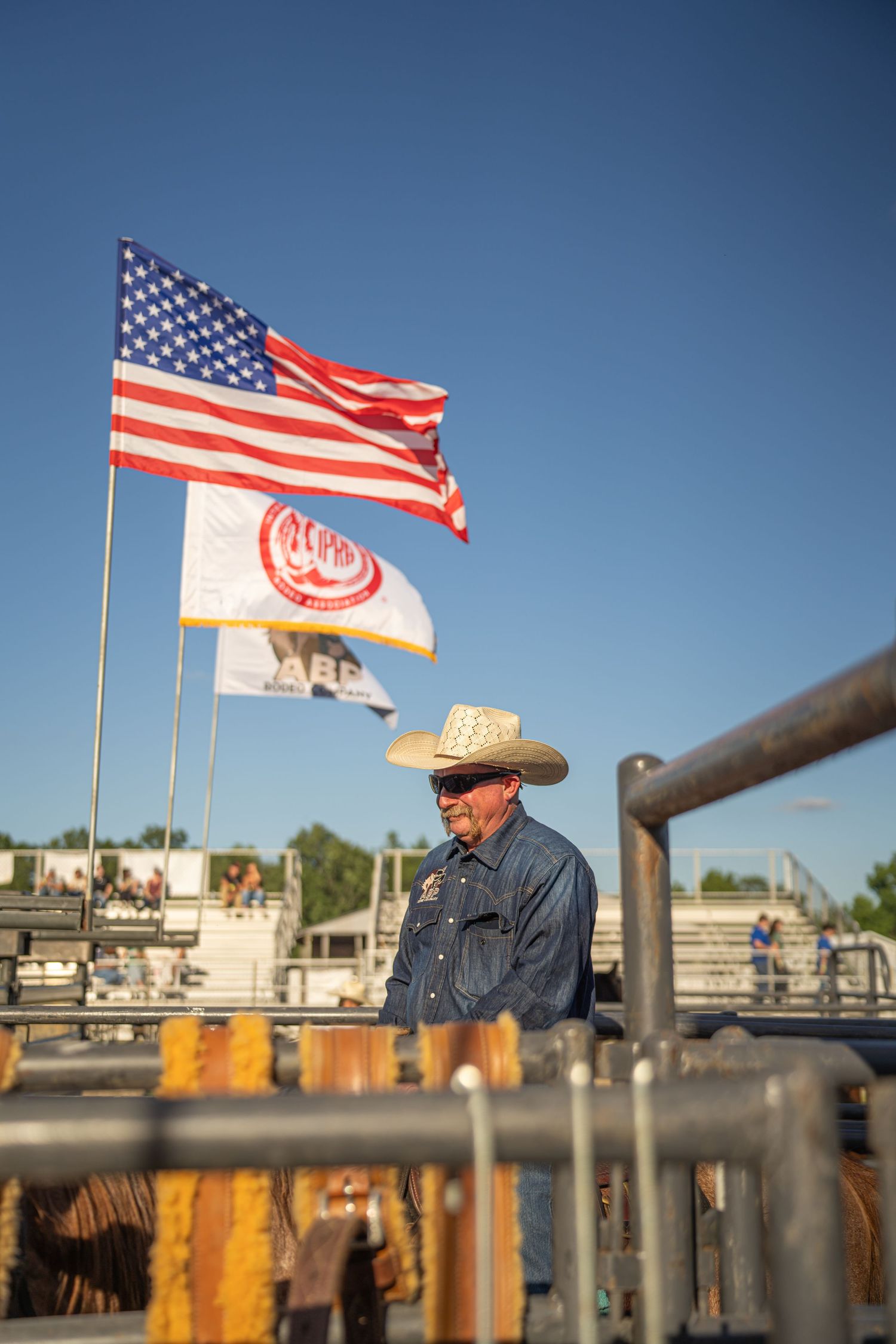 Cowboy in straw hat stands by metal gate with American and rodeo flags flying behind him.