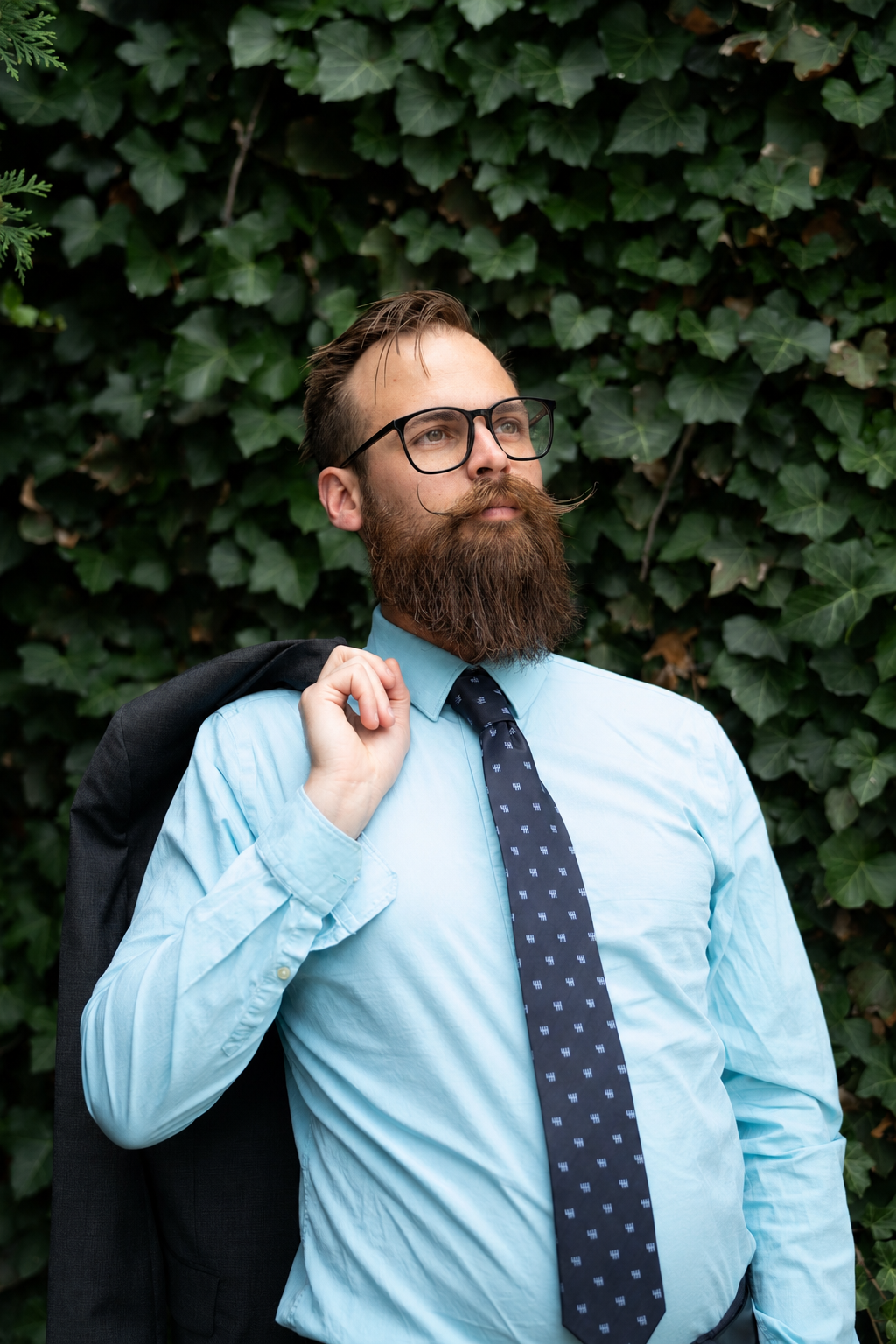 Bearded man in blue shirt, tie, and glasses holding jacket over shoulder against green ivy wall.
