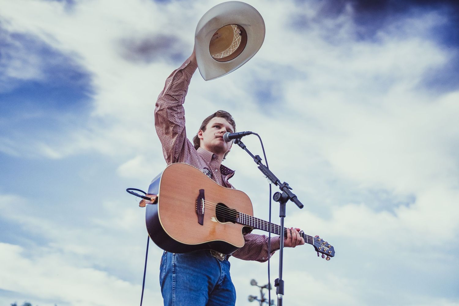 Country musician performing on stage, playing acoustic guitar and tipping cowboy hat to crowd under cloudy sky.