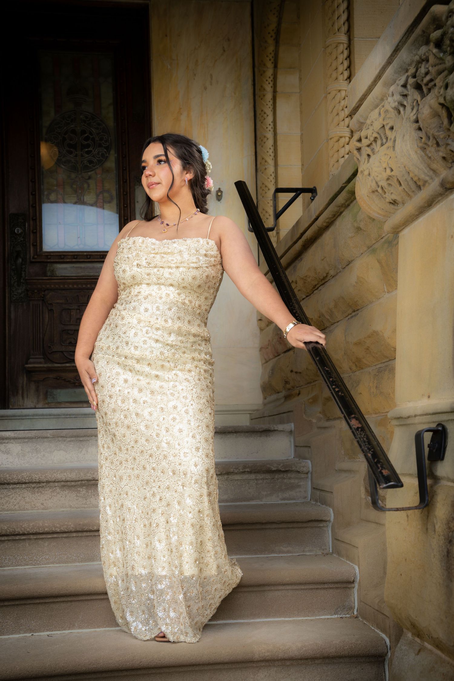 Woman in elegant gold sequin gown posing on stone steps beside ornate building entrance.