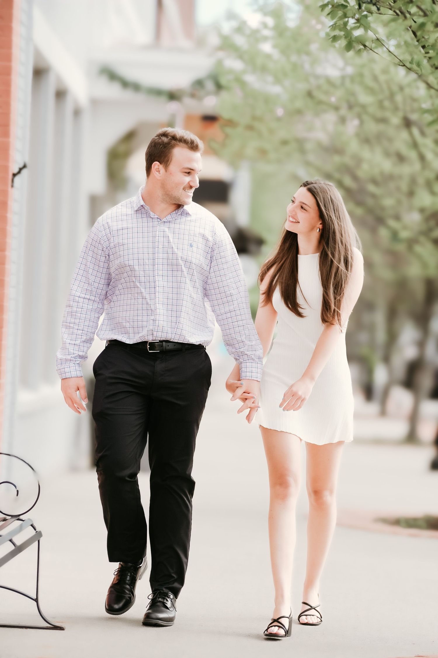 A happy couple walks hand in hand down a tree-lined sidewalk in casual elegant attire.