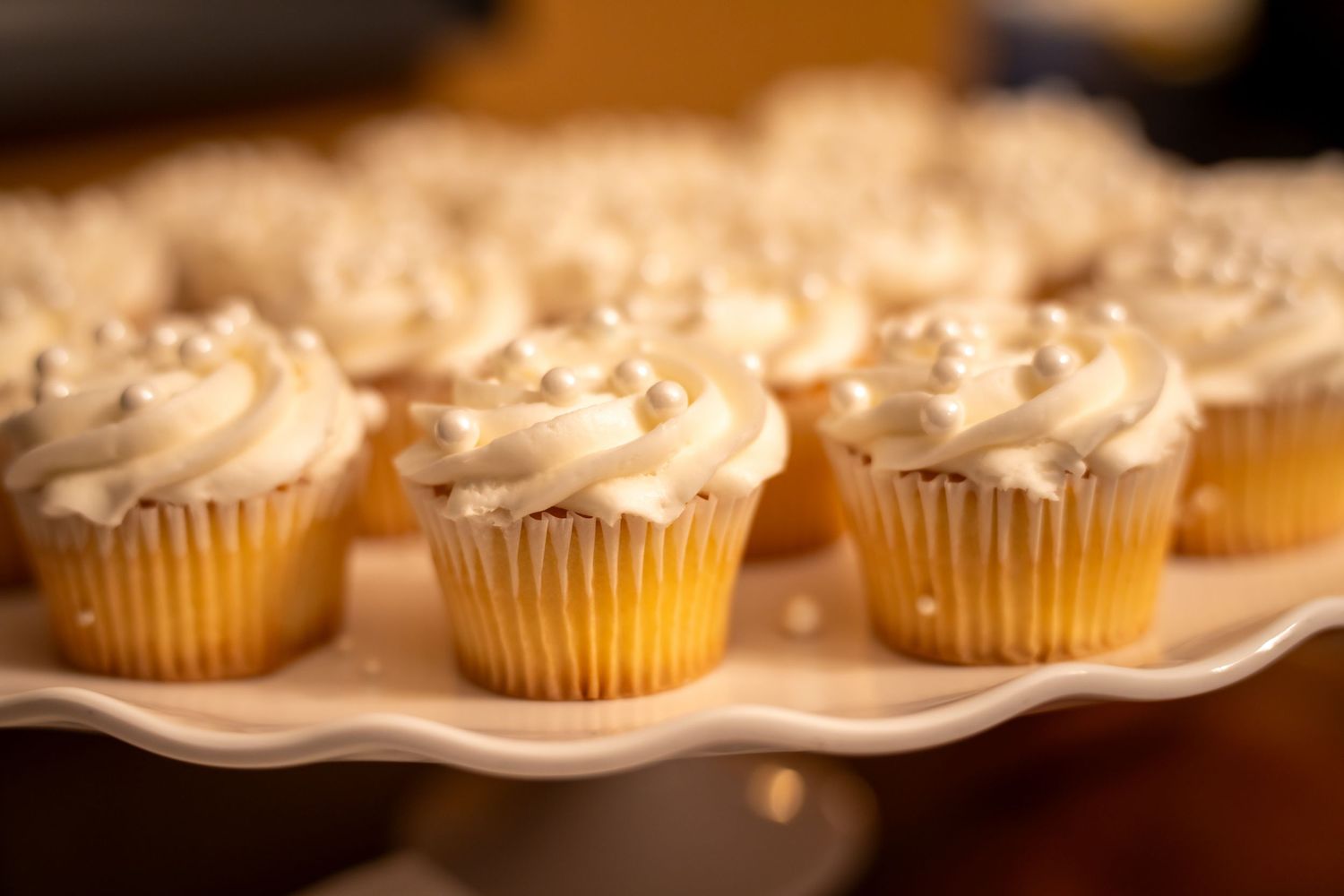 White frosted cupcakes with pearl sprinkles displayed on a white cake stand at a celebration.