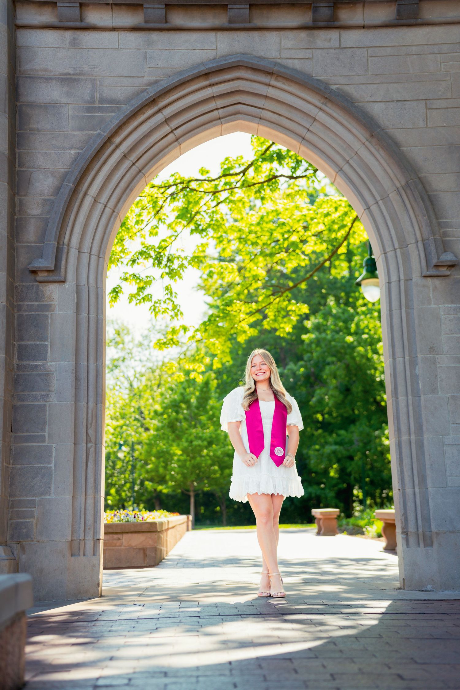 Smiling blonde graduate in white dress and pink stole posing under stone arch on sunny campus.