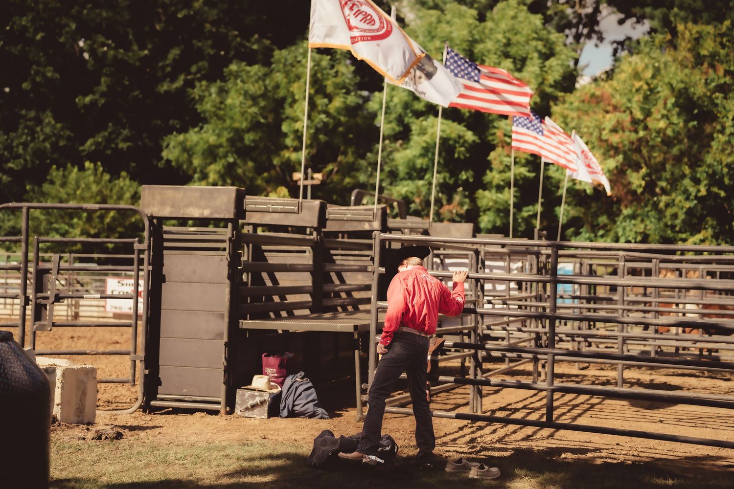 Rodeo arena with American flags flying above metal chutes and a worker in red shirt on sunny day.