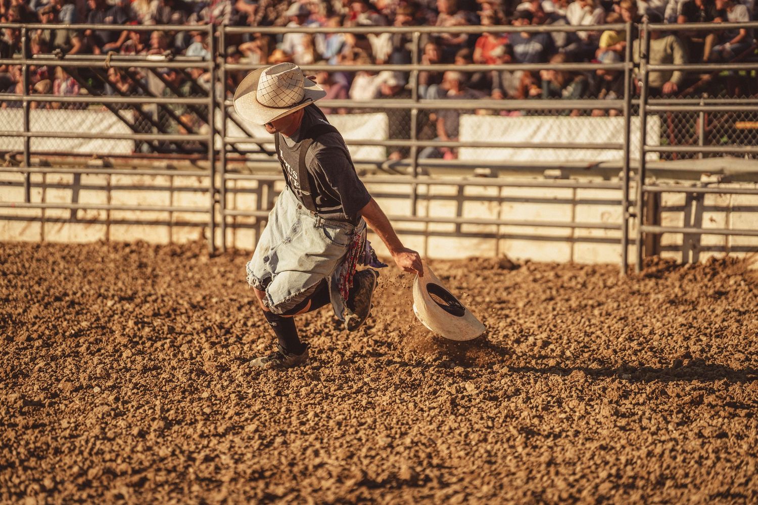 A cowboy kneels in the dirt of a rodeo arena picking up his hat after being thrown from a horse.