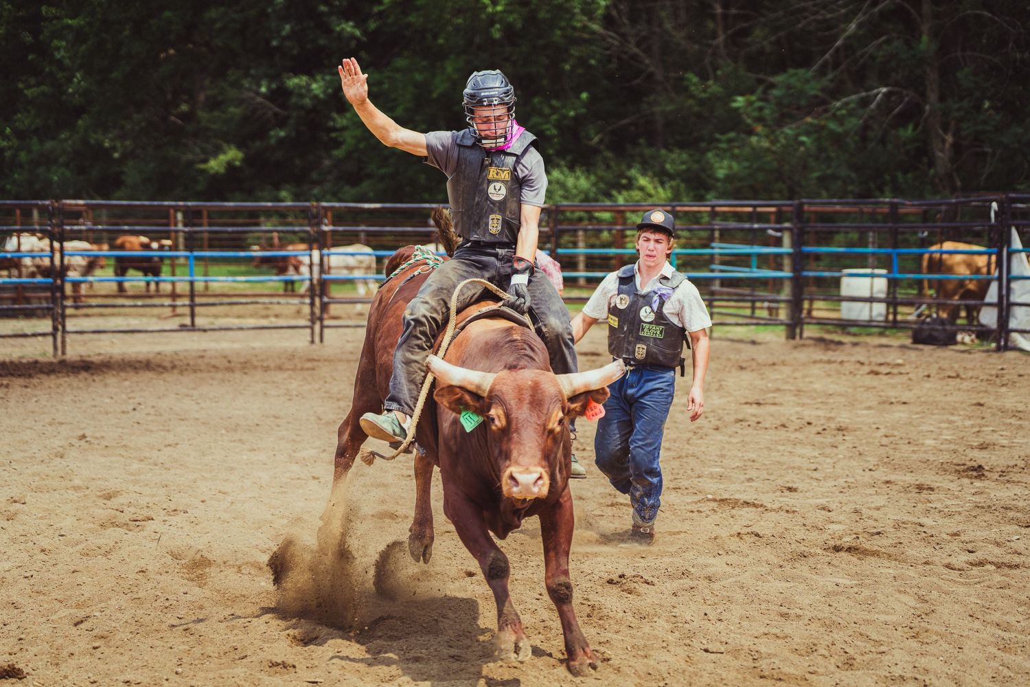 A cowboy rides a bucking bull at a rodeo while a rodeo clown stands nearby in a dirt arena.
