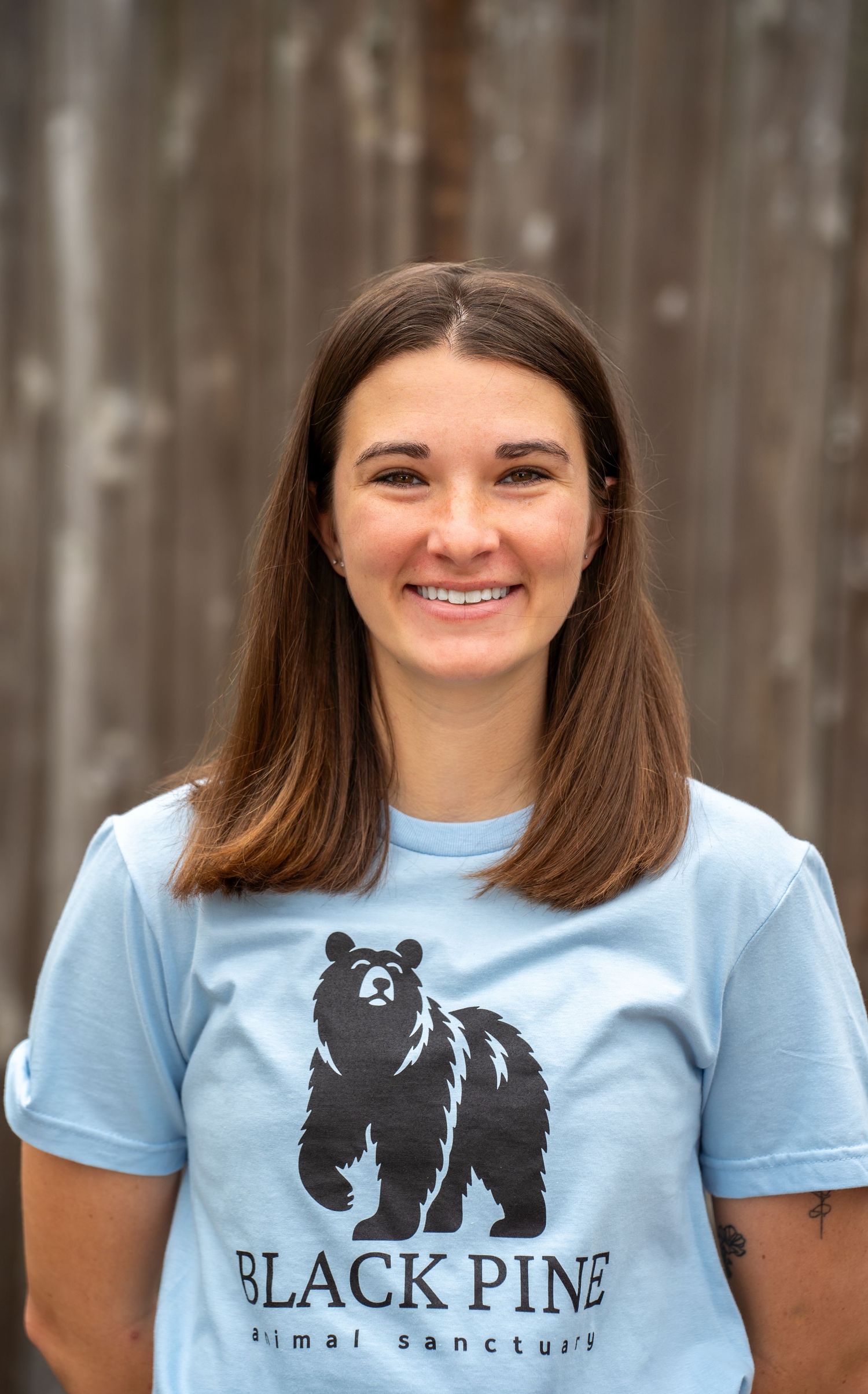 Smiling woman in a light blue Black Pine Animal Sanctuary t-shirt posing outdoors against a wooden backdrop.