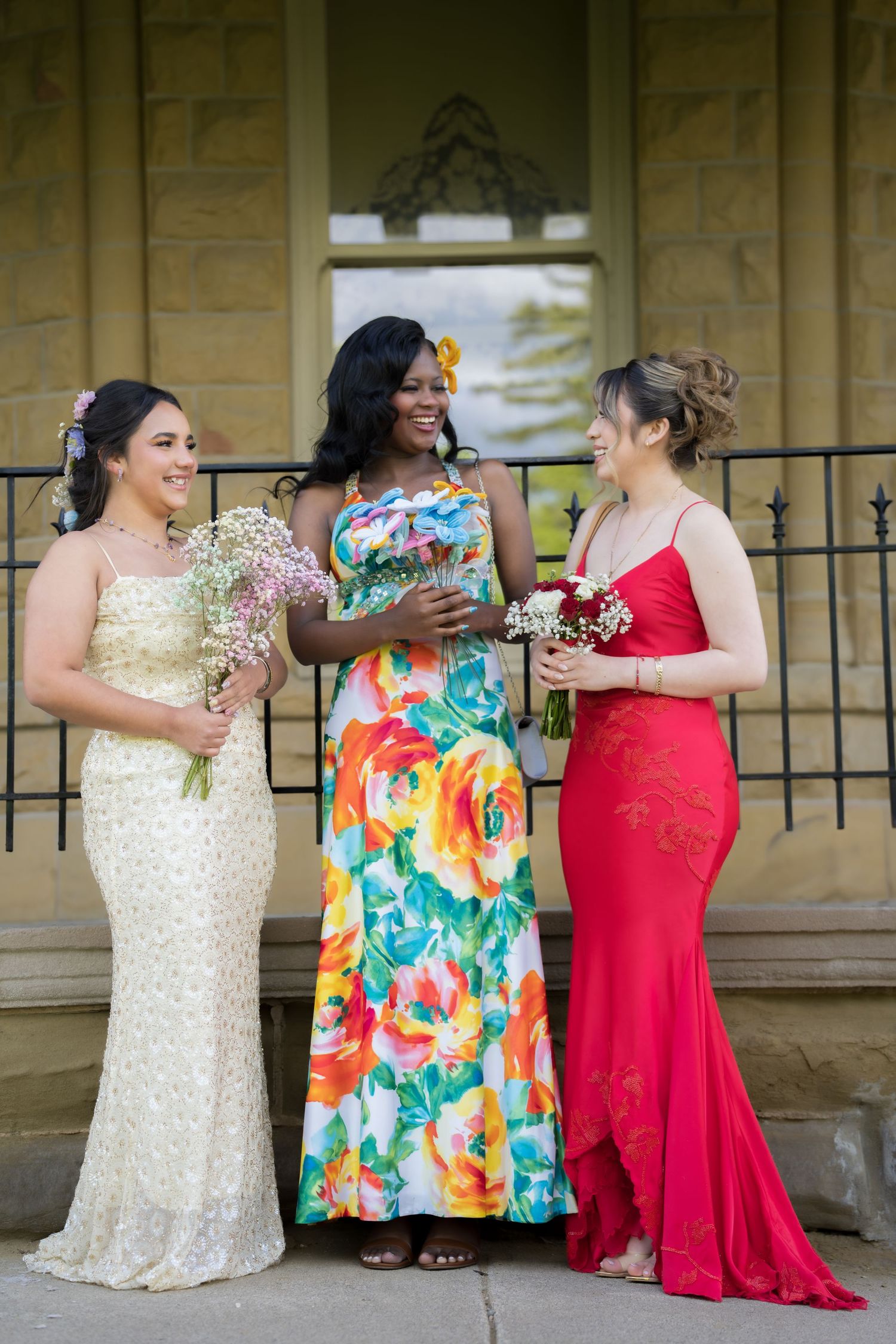 Three women in elegant gowns holding bouquets pose together on stone steps at a formal outdoor event.