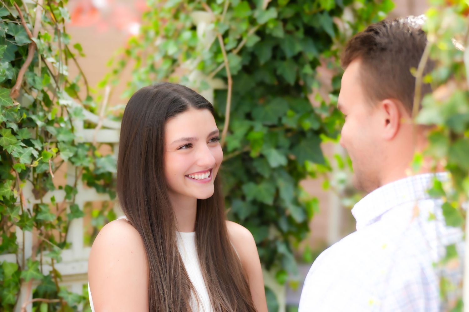 A young woman with long brown hair smiles at a man outdoors near a green ivy backdrop.