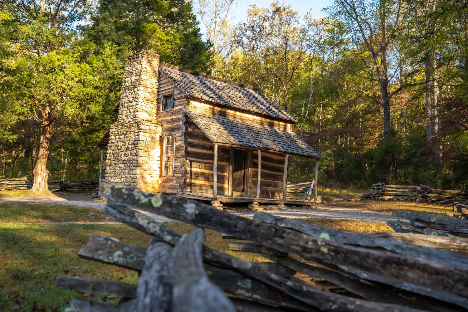 Historic log cabin with stone chimney surrounded by autumn trees and split-rail fence in golden light.