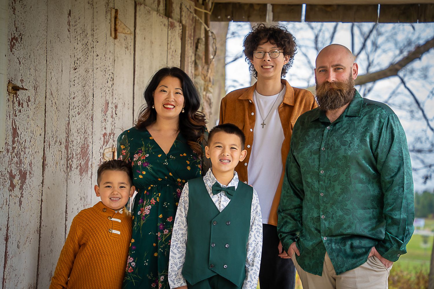 A family of five poses together smiling in front of a rustic white wooden barn wall.