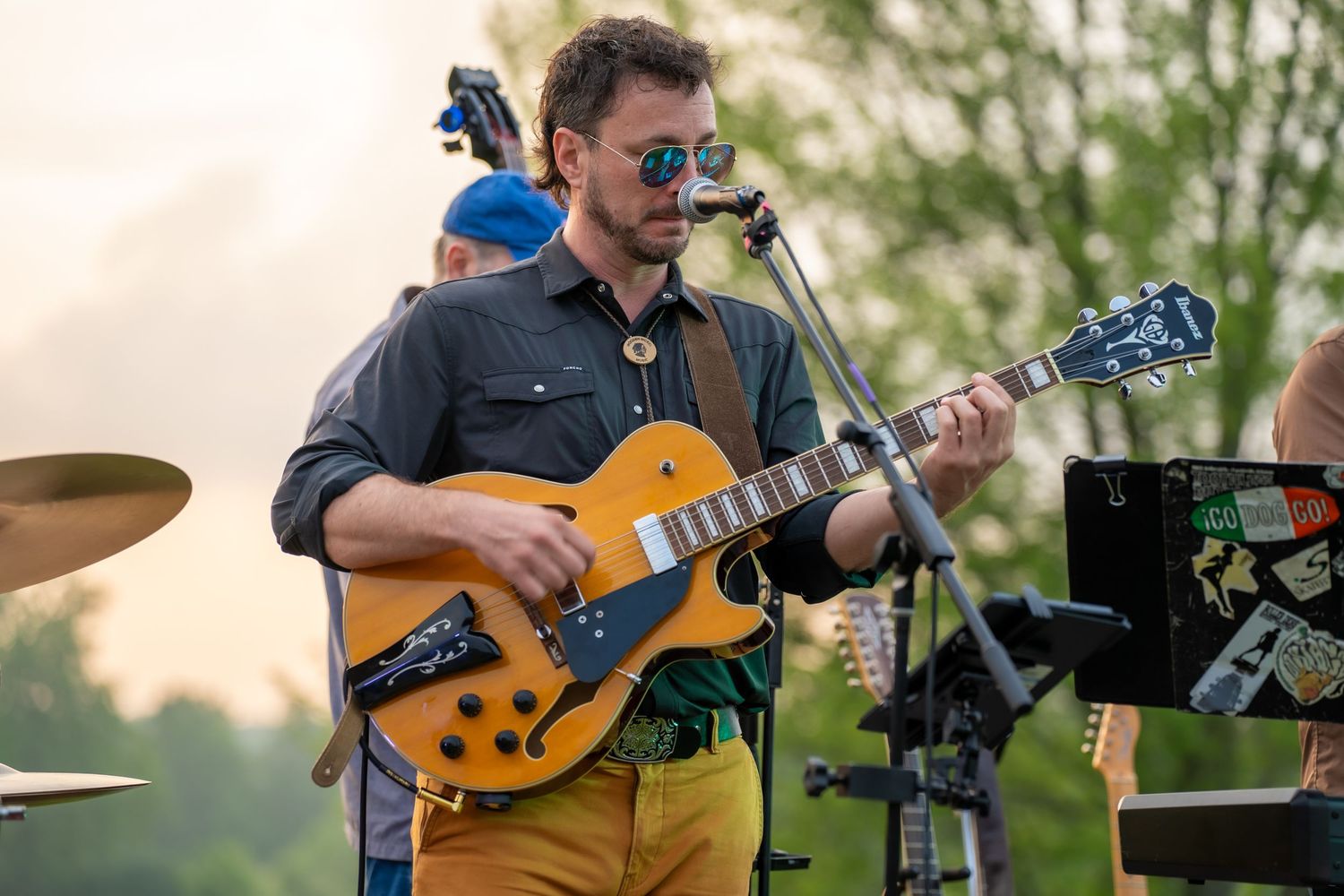 Male guitarist playing archtop electric guitar at outdoor music festival wearing sunglasses.