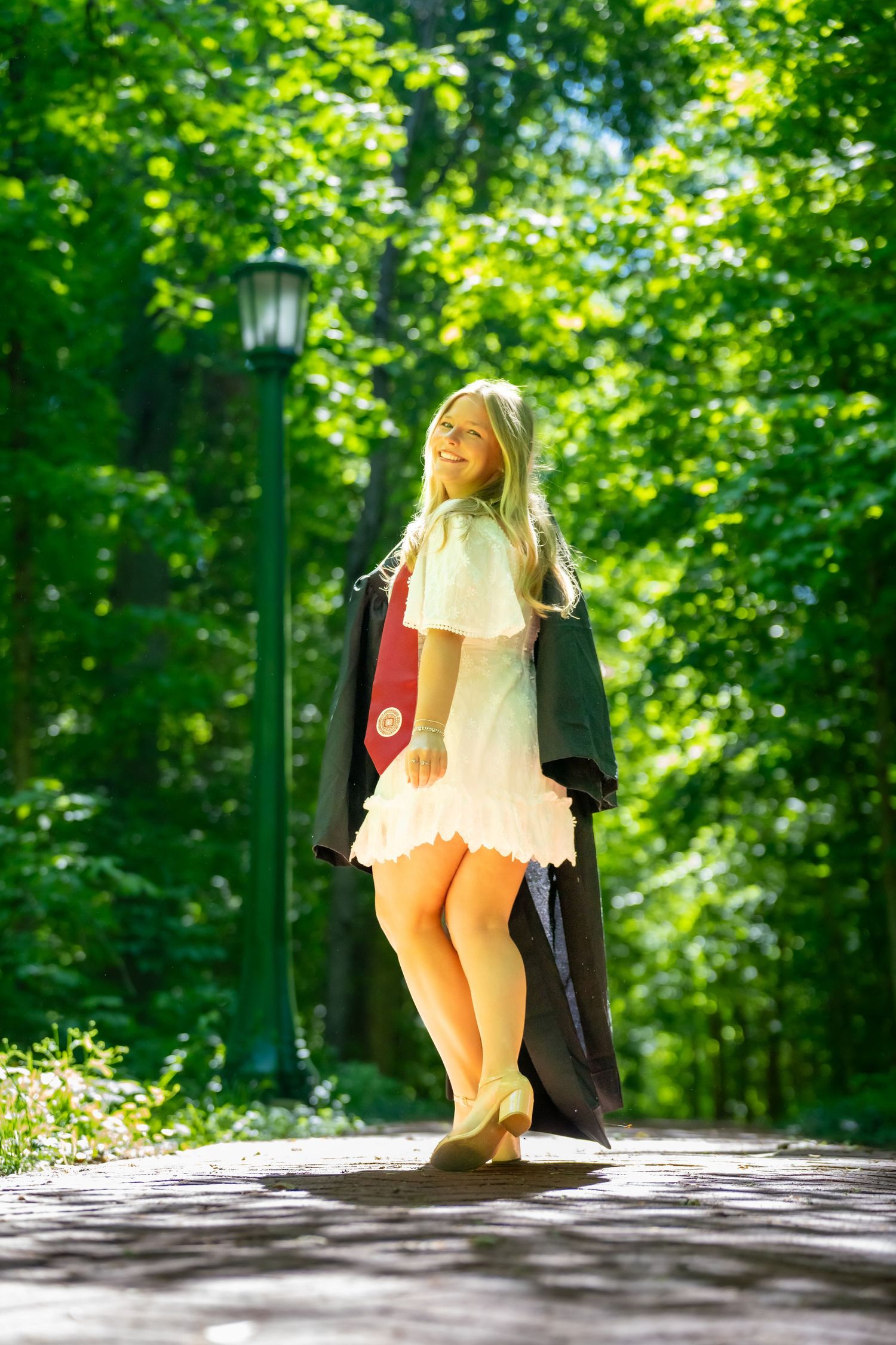 Blonde graduate in white dress carrying black graduation gown on sunny tree-lined campus path.