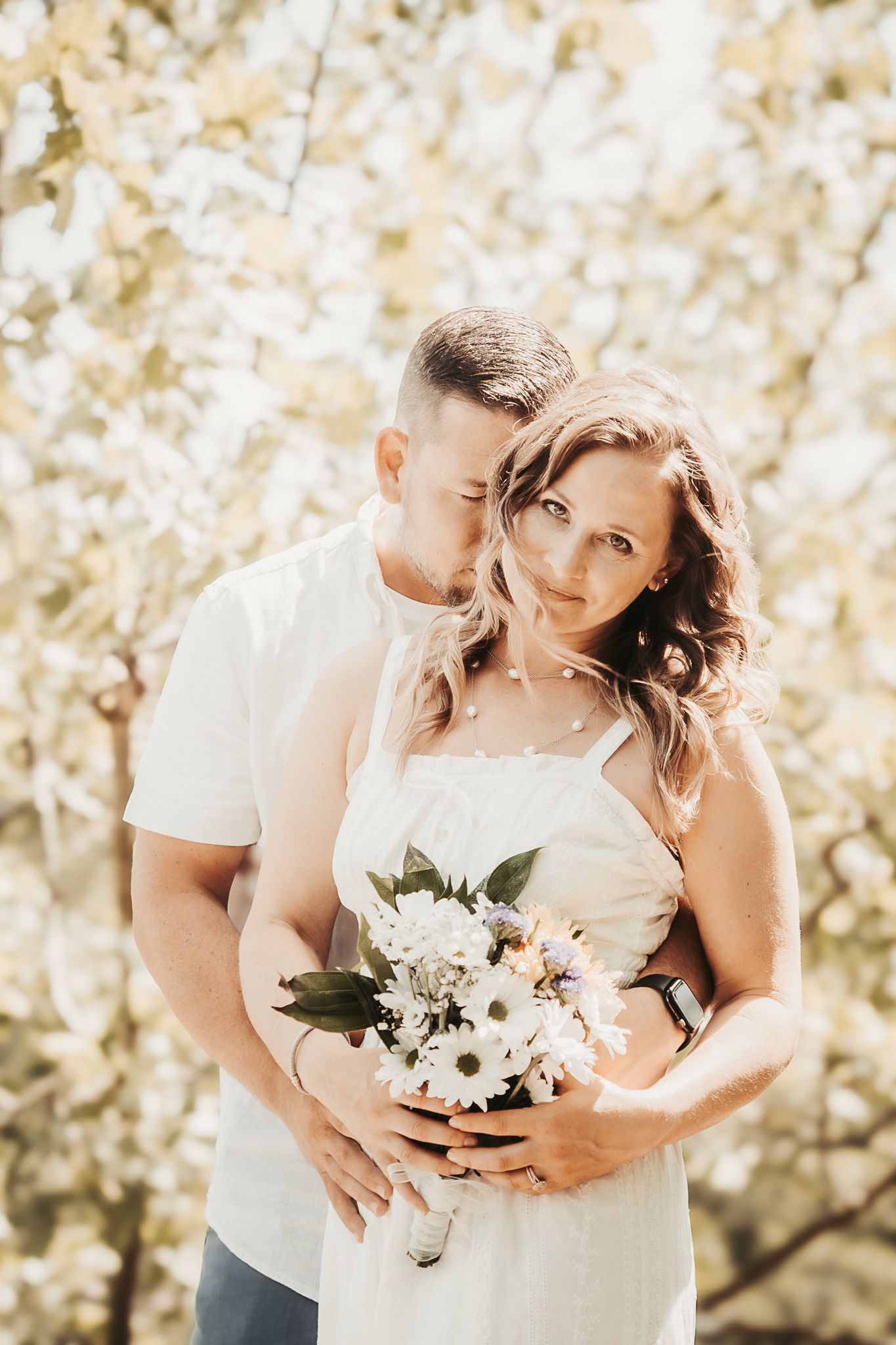 Romantic couple embracing outdoors with man holding woman from behind, she holds white flowers.