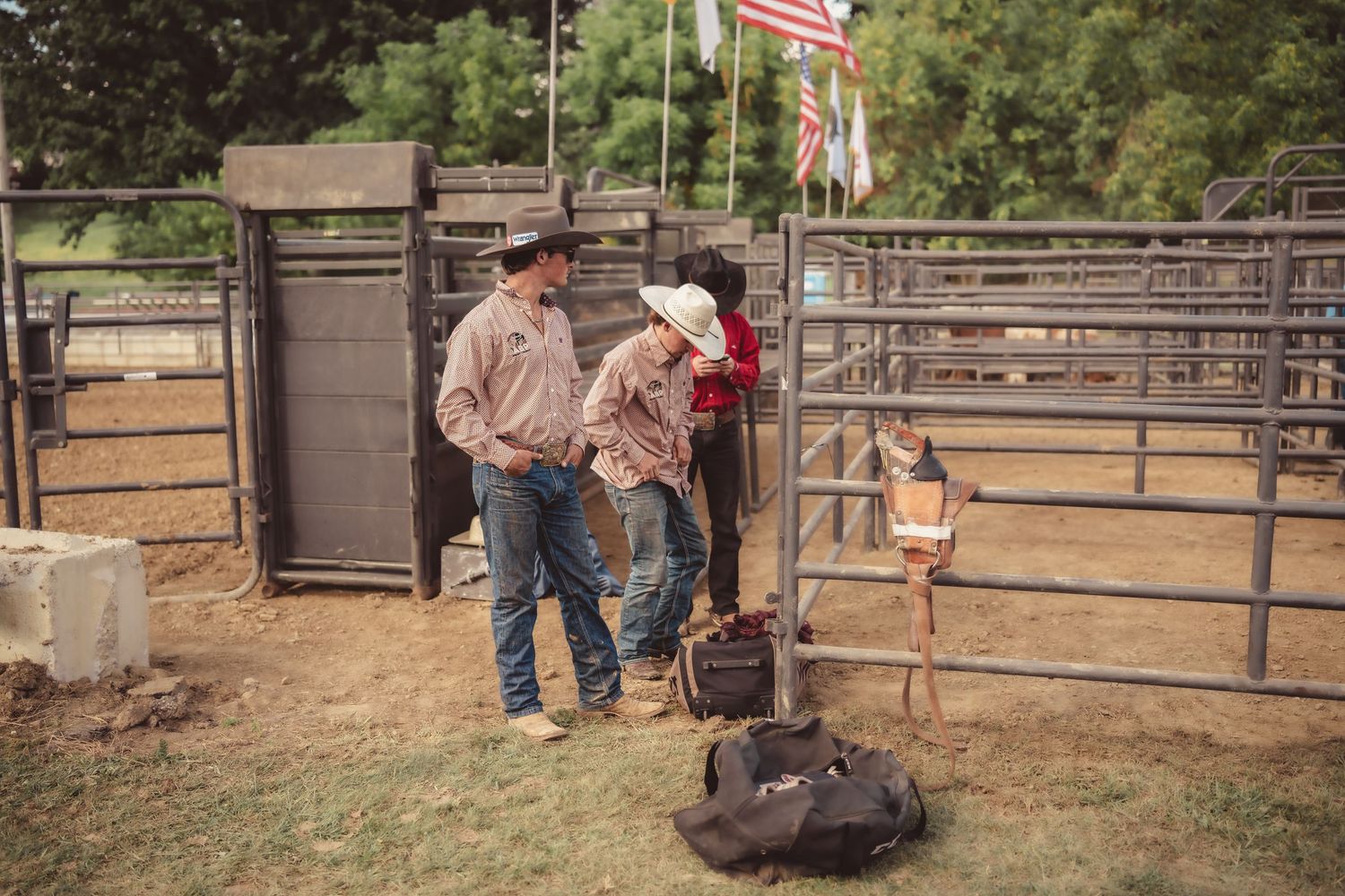 Two cowboys in western wear standing near metal rodeo chutes with American flag and gear bags.