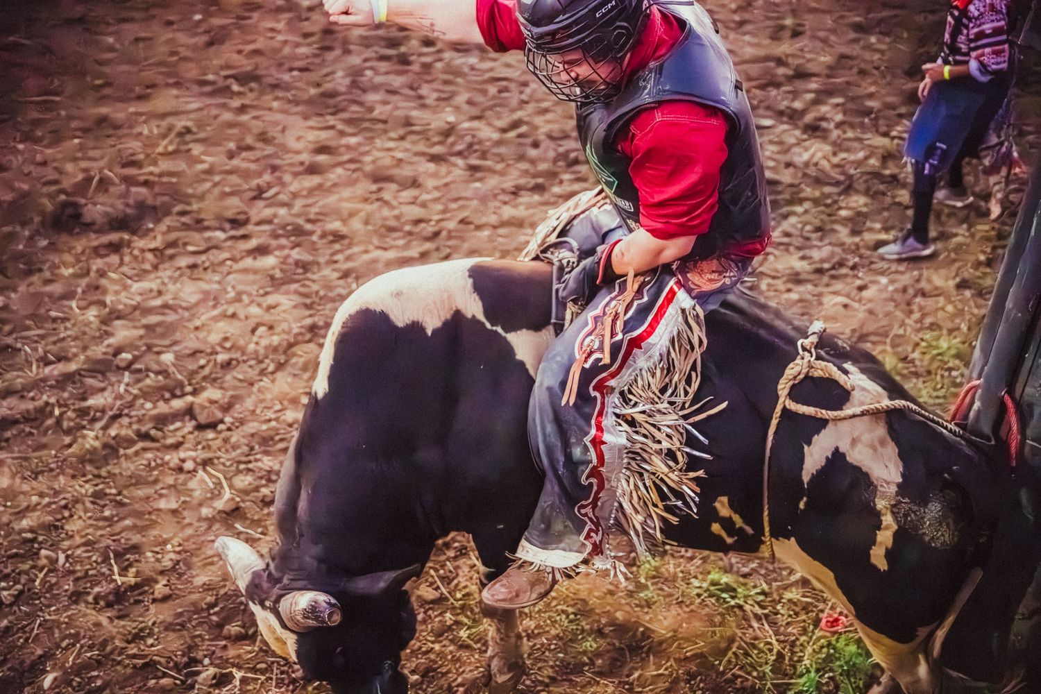 A bull rider in red vest clings to a black and white bull during an intense rodeo competition outdoors.