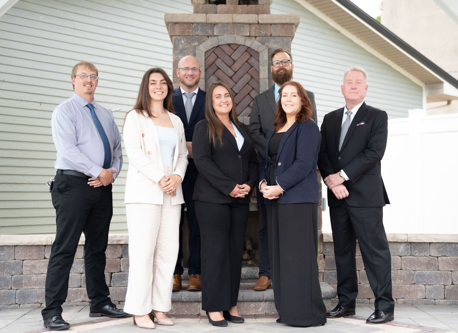 Group of seven business professionals posing together outdoors in front of a decorative stone pillar.