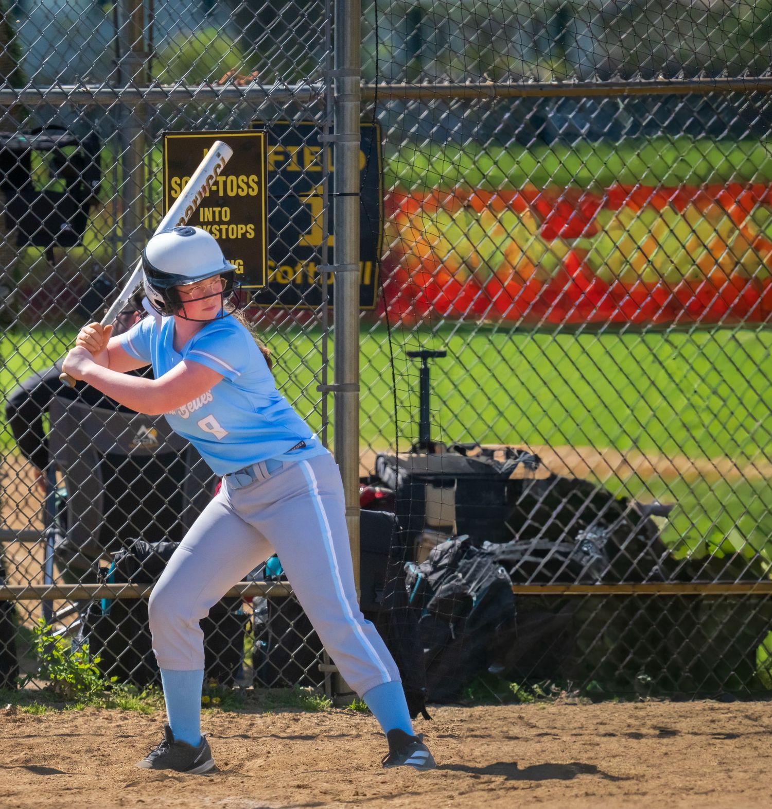 Female softball player in blue uniform stands ready at bat on a sunny day near a chain-link fence.