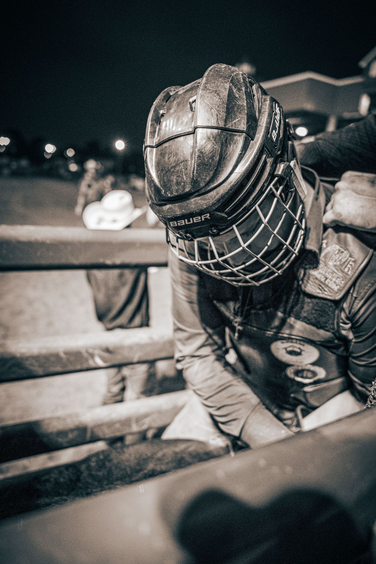 Hockey player in worn Bauer helmet with cage leans over boards during a night game.
