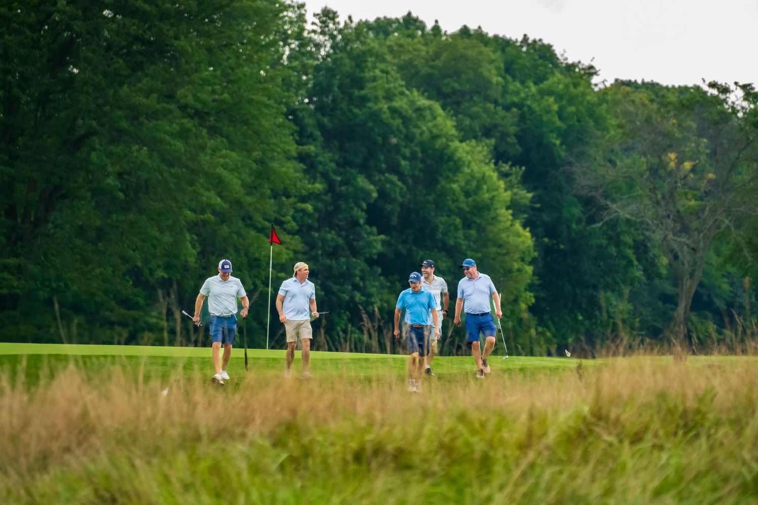 Group of golfers walking across a green fairway surrounded by dense trees on a golf course.