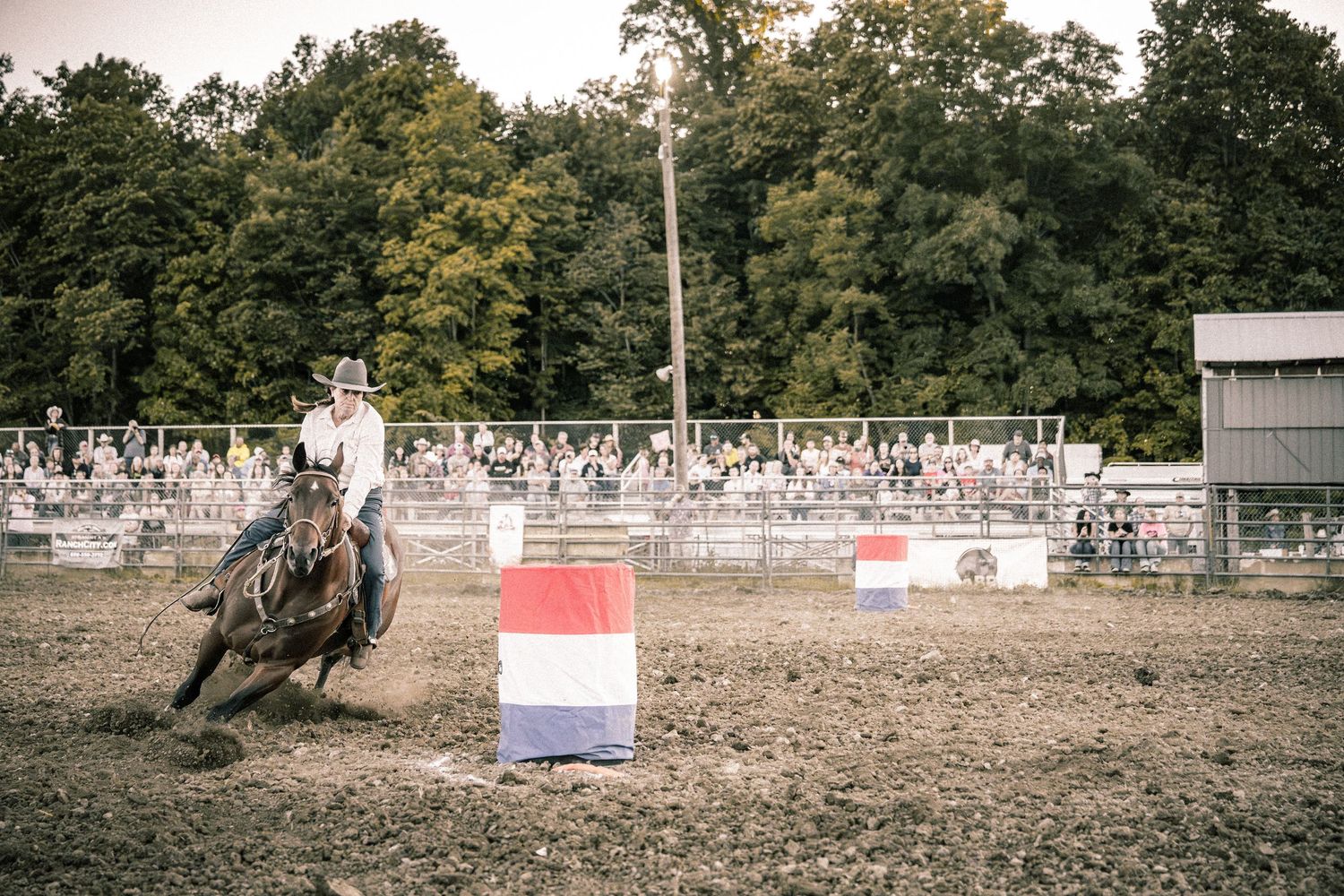 Cowboy on horseback navigating around a red, white, and blue barrel during a rodeo competition with spectators watching.