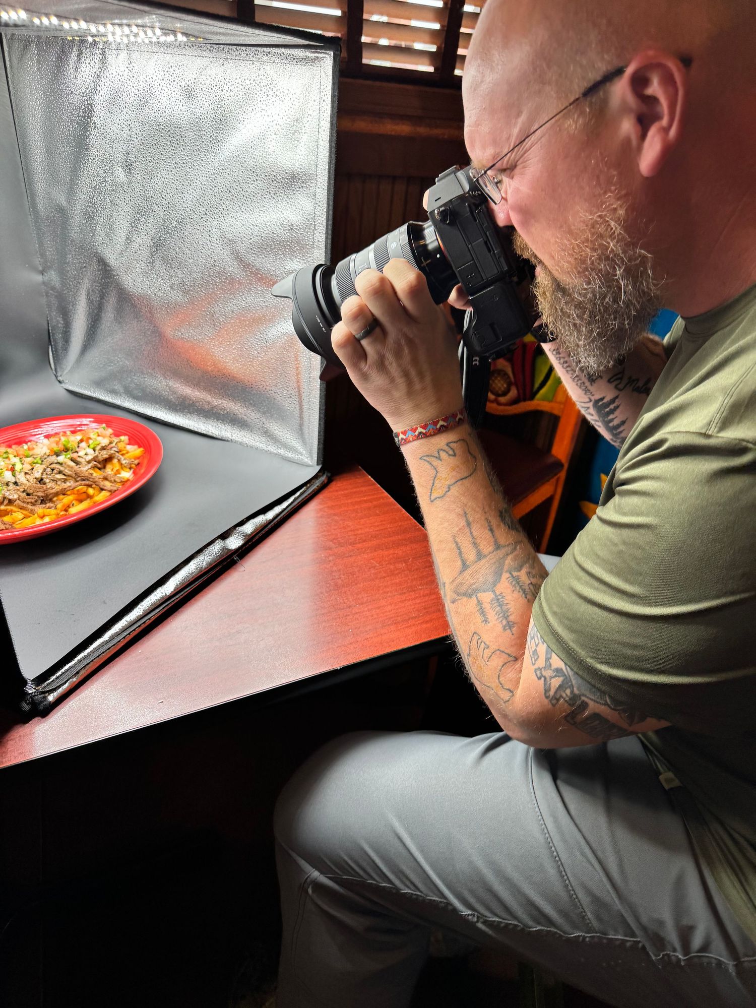 Tattooed bald man with beard photographing food on red plate using DSLR camera in a portable photo studio lightbox.