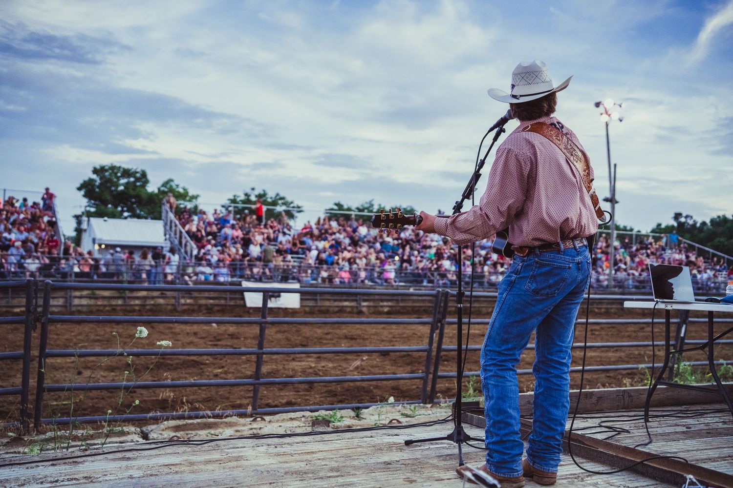 Cowboy performer in white hat addresses large rodeo crowd from stage at outdoor country western event.