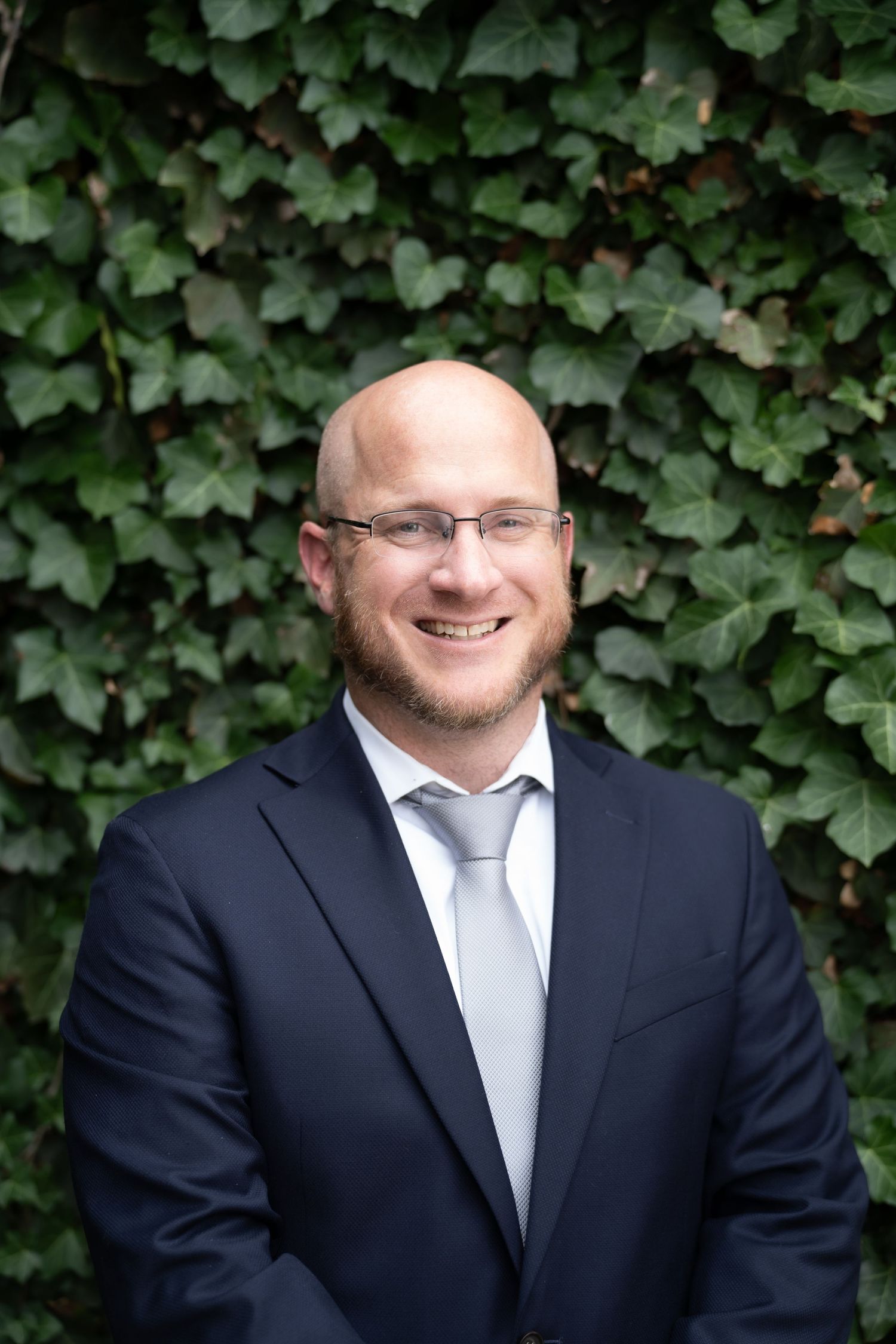 Smiling bald man with glasses in a dark suit and gray tie, posing in front of a lush green ivy wall.