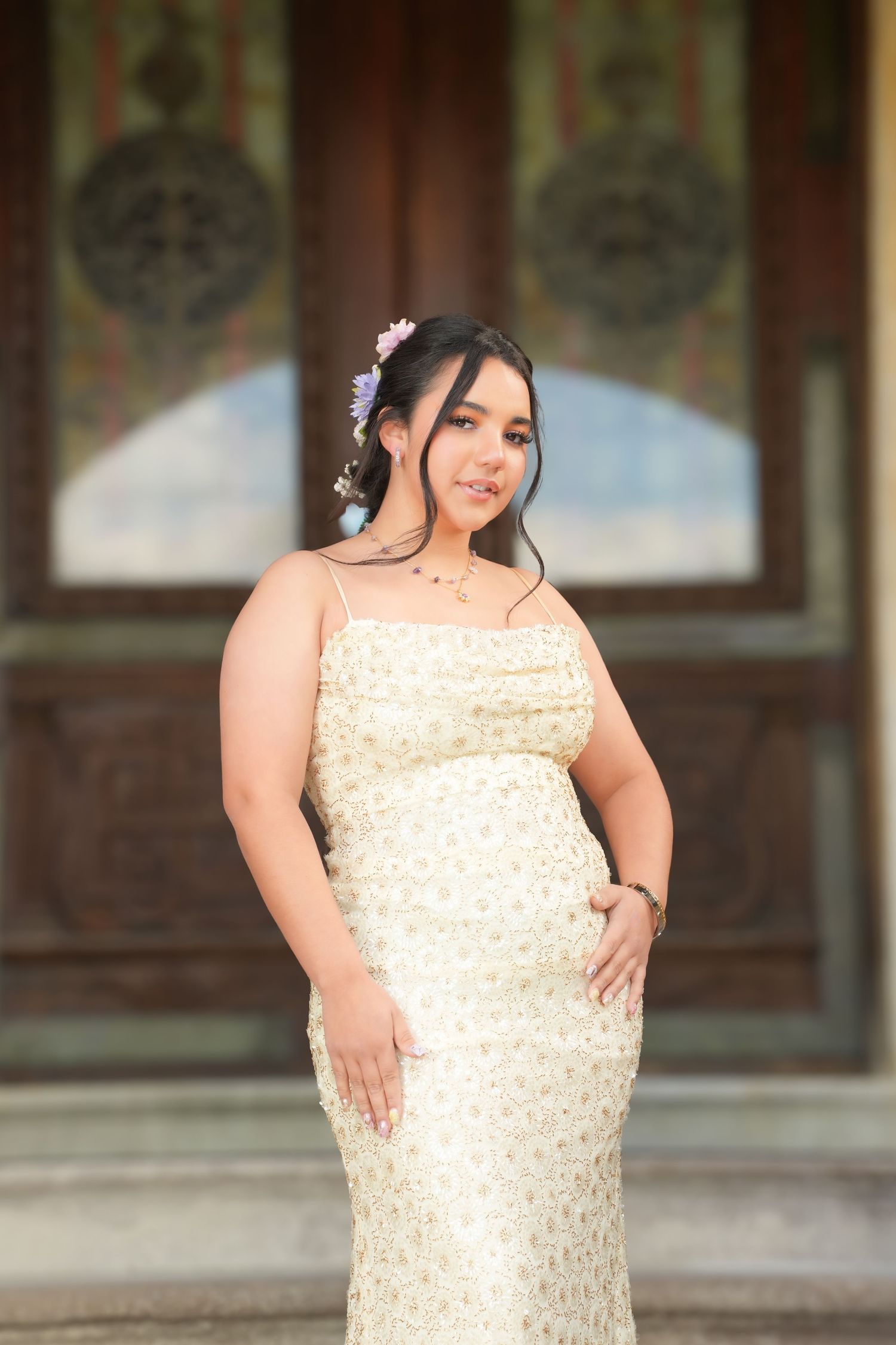 Young woman in floral cream spaghetti strap gown with purple hair flowers poses before ornate wooden doors.