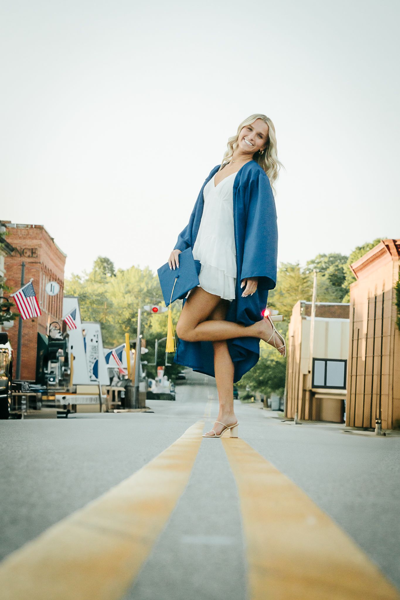 Joyful blonde woman in blue graduation gown posing on yellow street lines in small town.