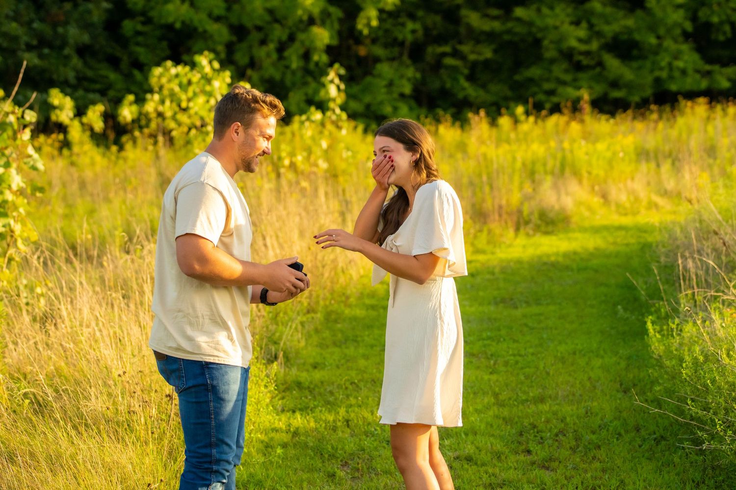 Man placing engagement ring on laughing woman's finger on a sunny green meadow path.