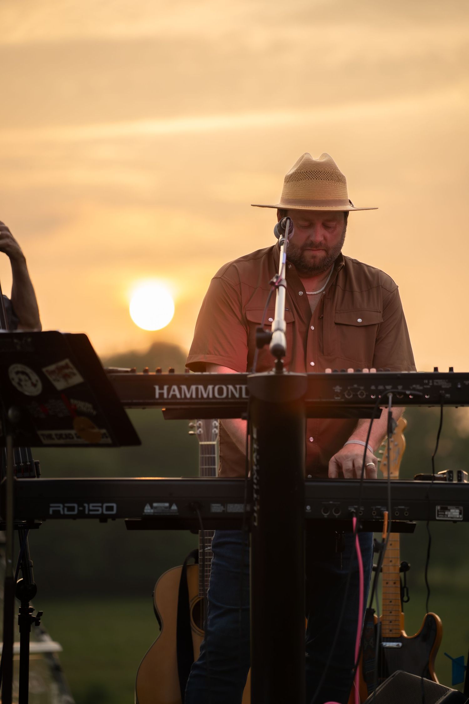 Male musician in a wide-brim hat playing a Hammond keyboard on stage during a vibrant outdoor sunset performance.