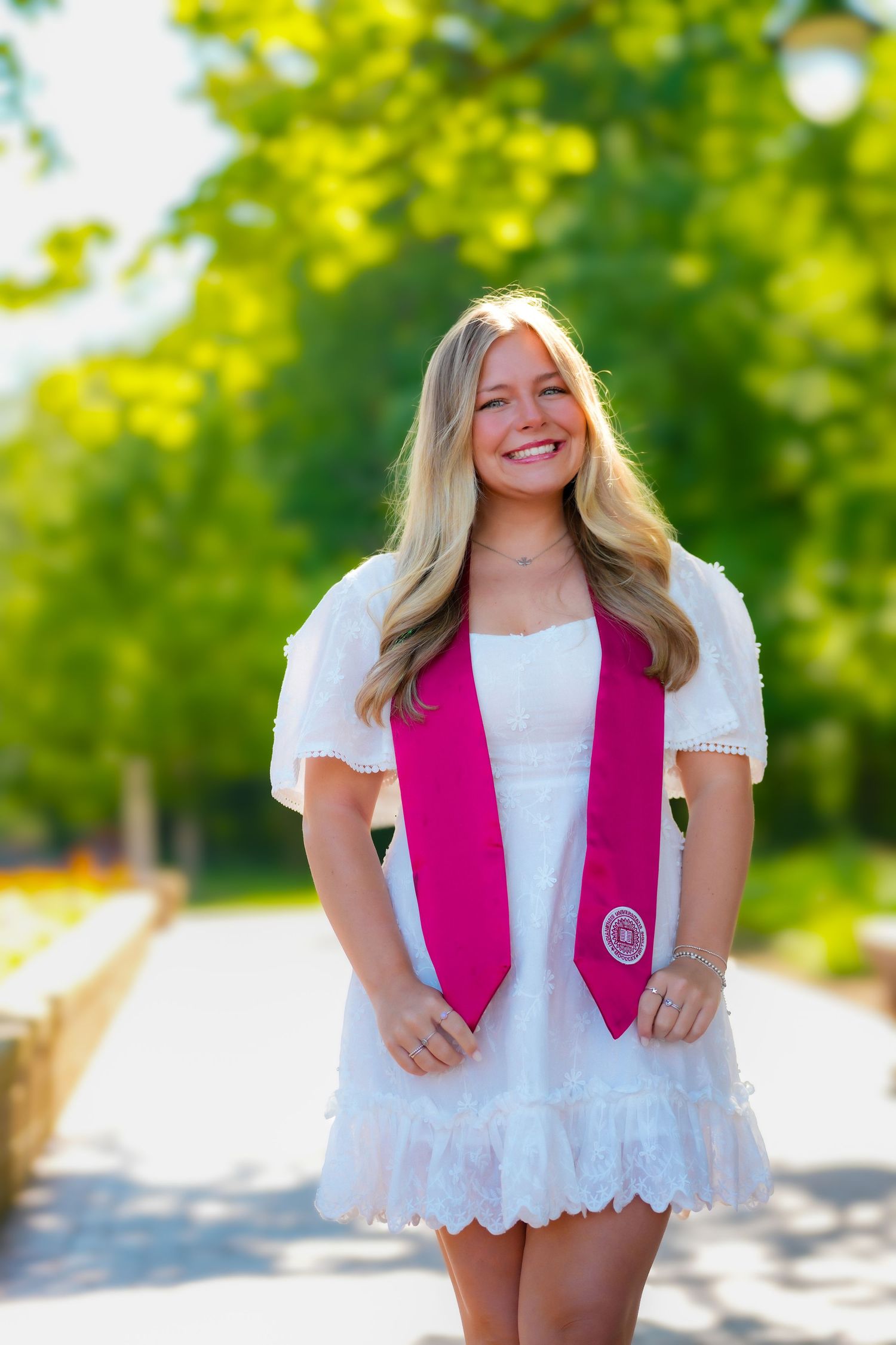 Happy blonde graduate in white dress wearing pink Indiana University graduation stole on sunny path.