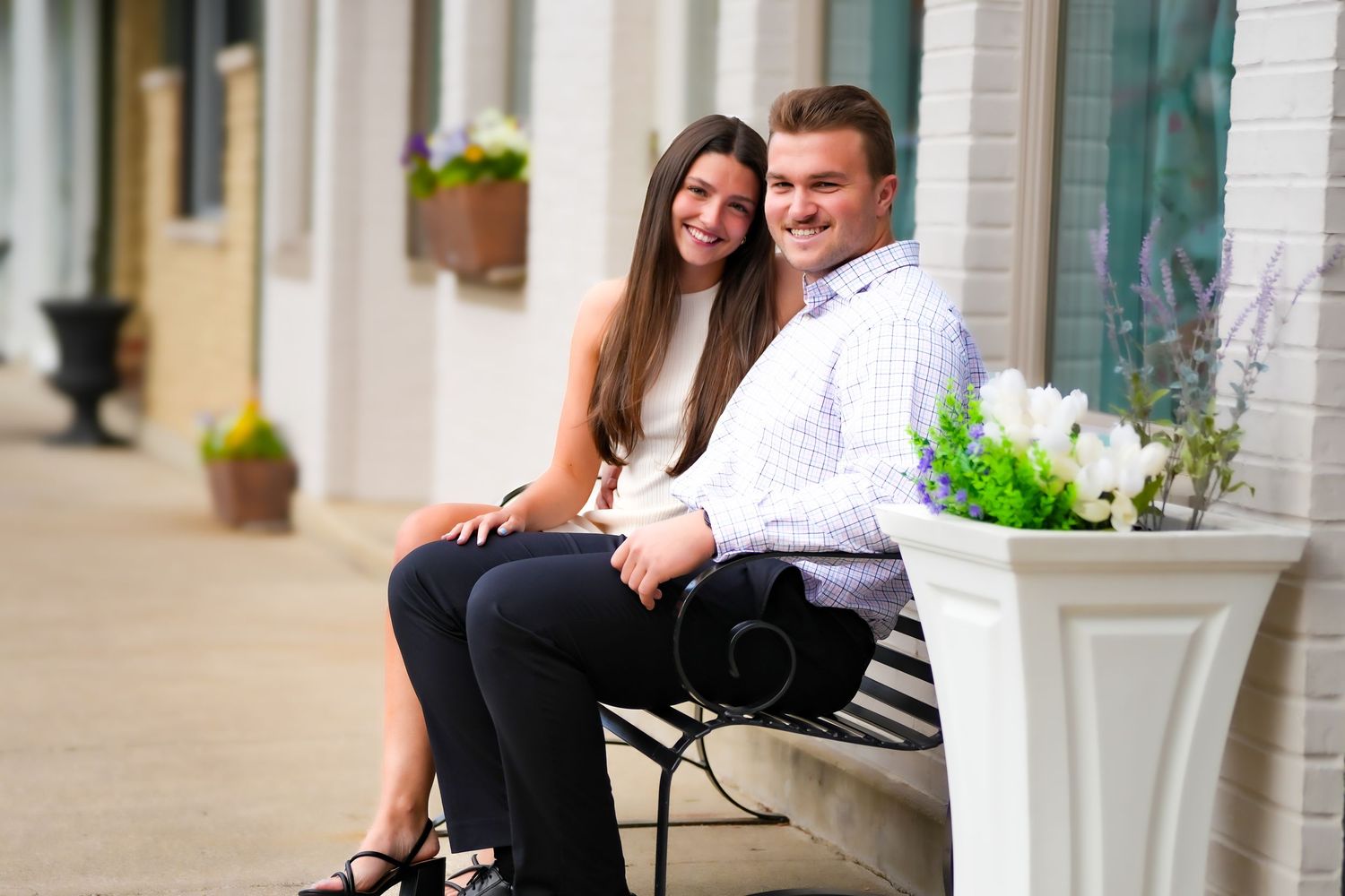 A smiling couple sits together on a bench outside a building with flower planters nearby.