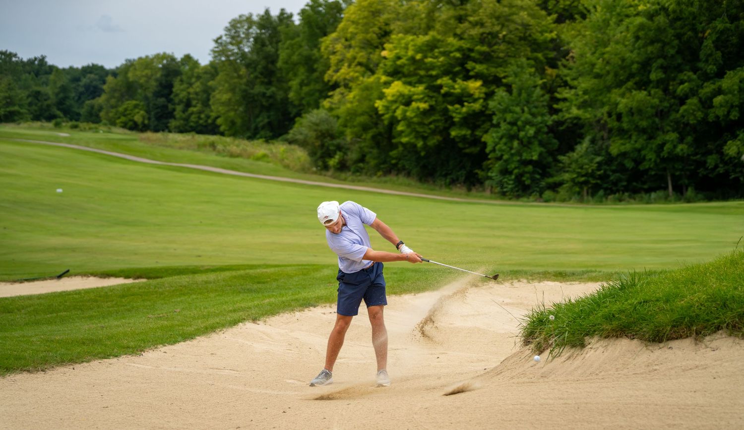 Golfer hitting a shot from a sand bunker on a lush green golf course surrounded by trees.