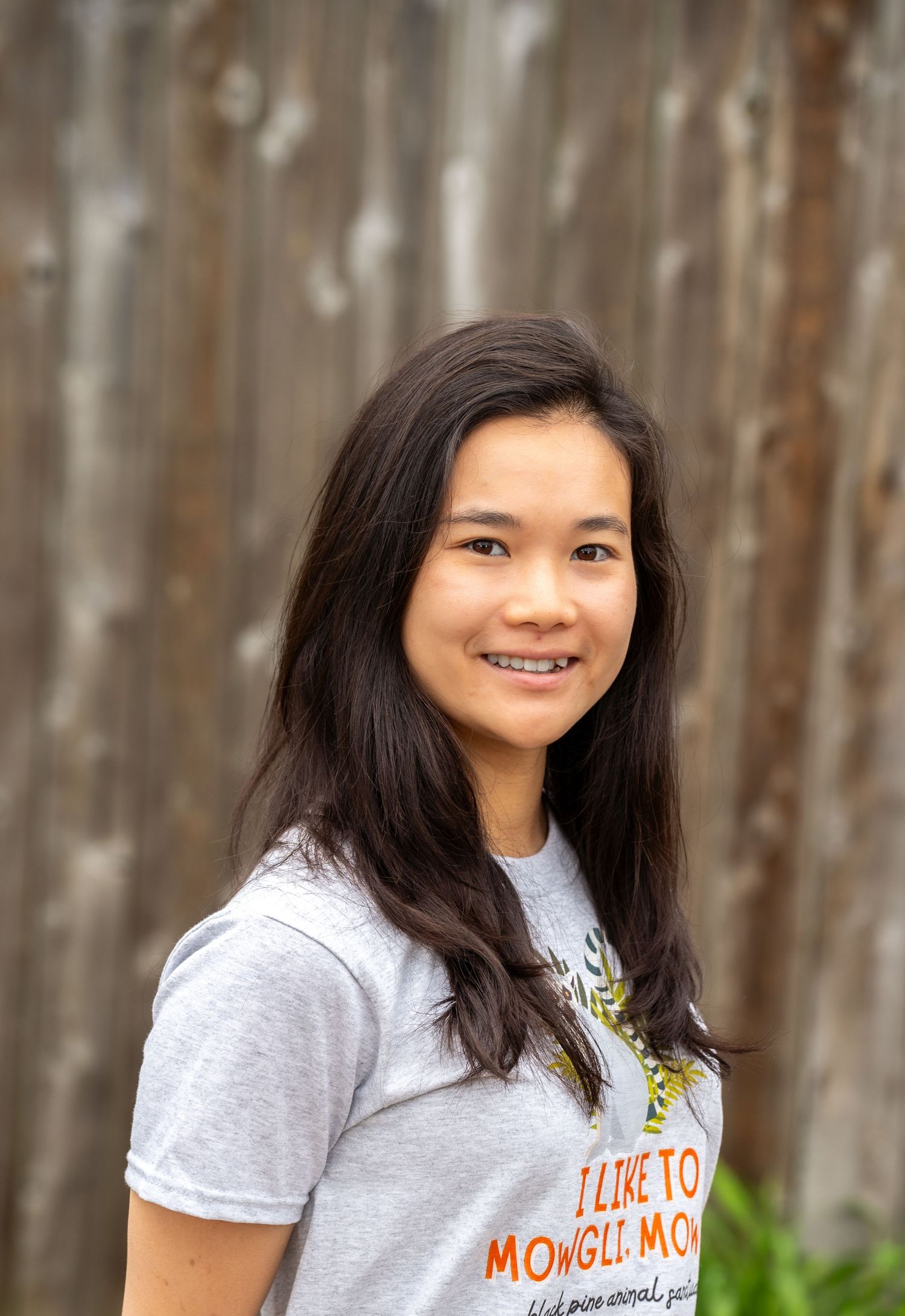 Young woman with long dark hair smiling outdoors, wearing a gray Mowgli-themed t-shirt against a wooden backdrop.
