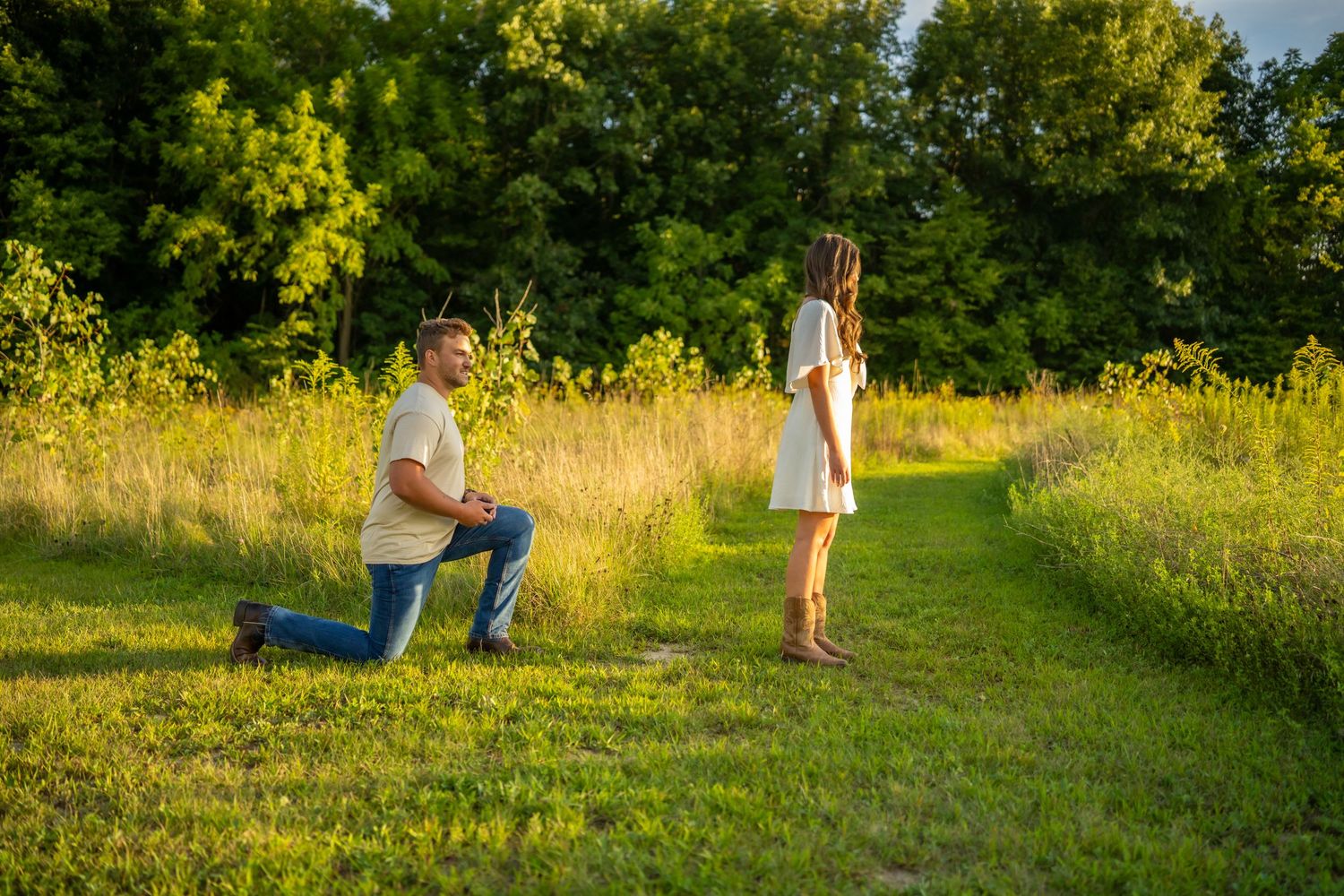 A man kneels on one knee proposing to a woman on a sunny green meadow path.