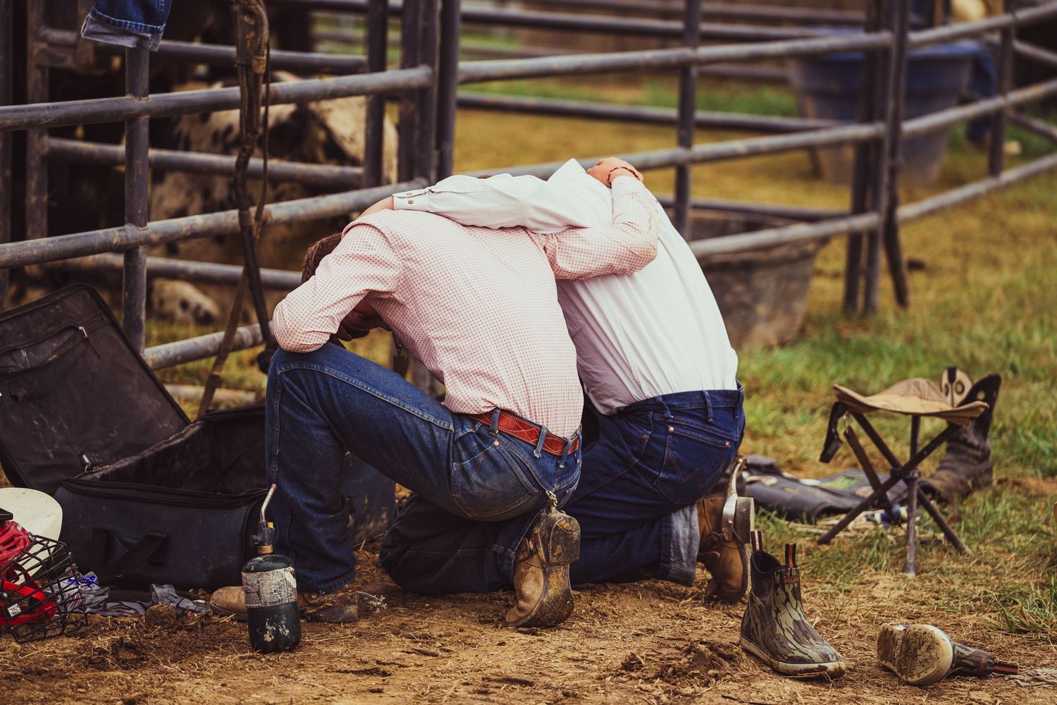 Two cowboys kneel together in prayer at a rodeo arena, wearing pink shirts and jeans with boots.
