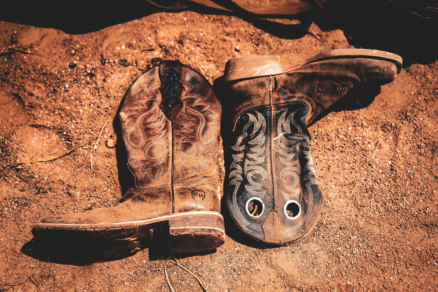 Worn brown leather cowboy boots resting on red dirt ground, showcasing rustic western style and aged craftsmanship.