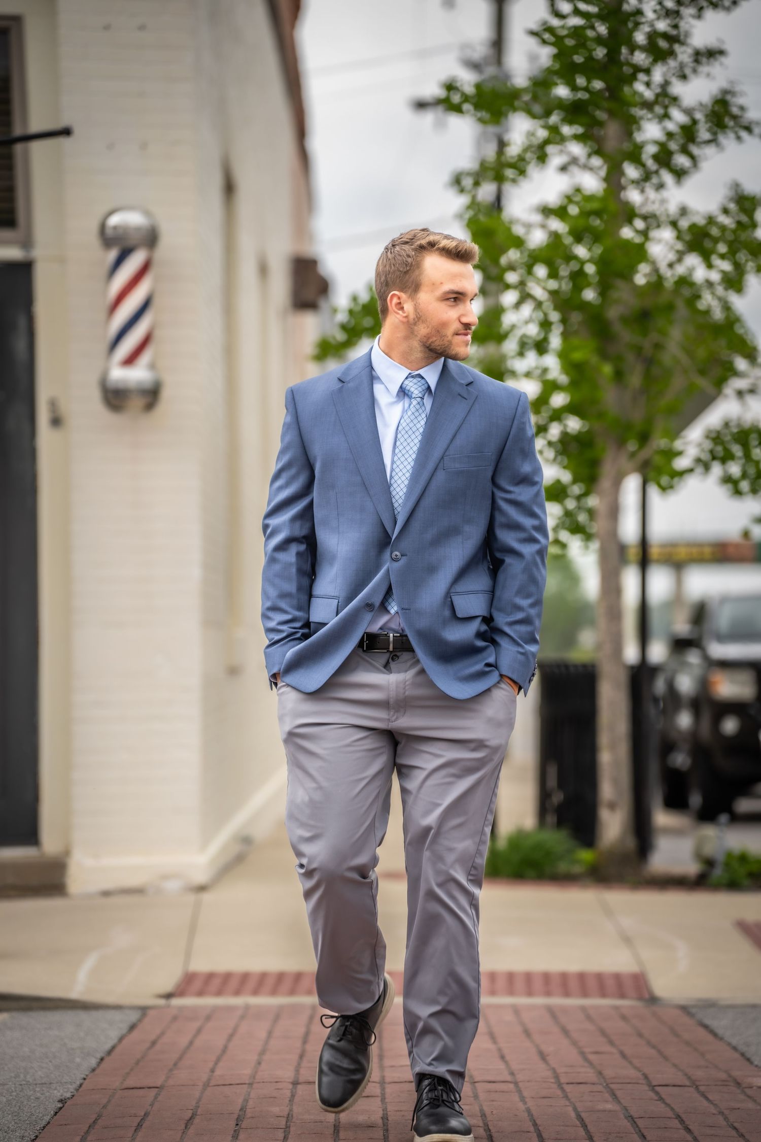 A well-dressed man in a blue blazer and gray pants strolls past a classic barber pole on a sidewalk.