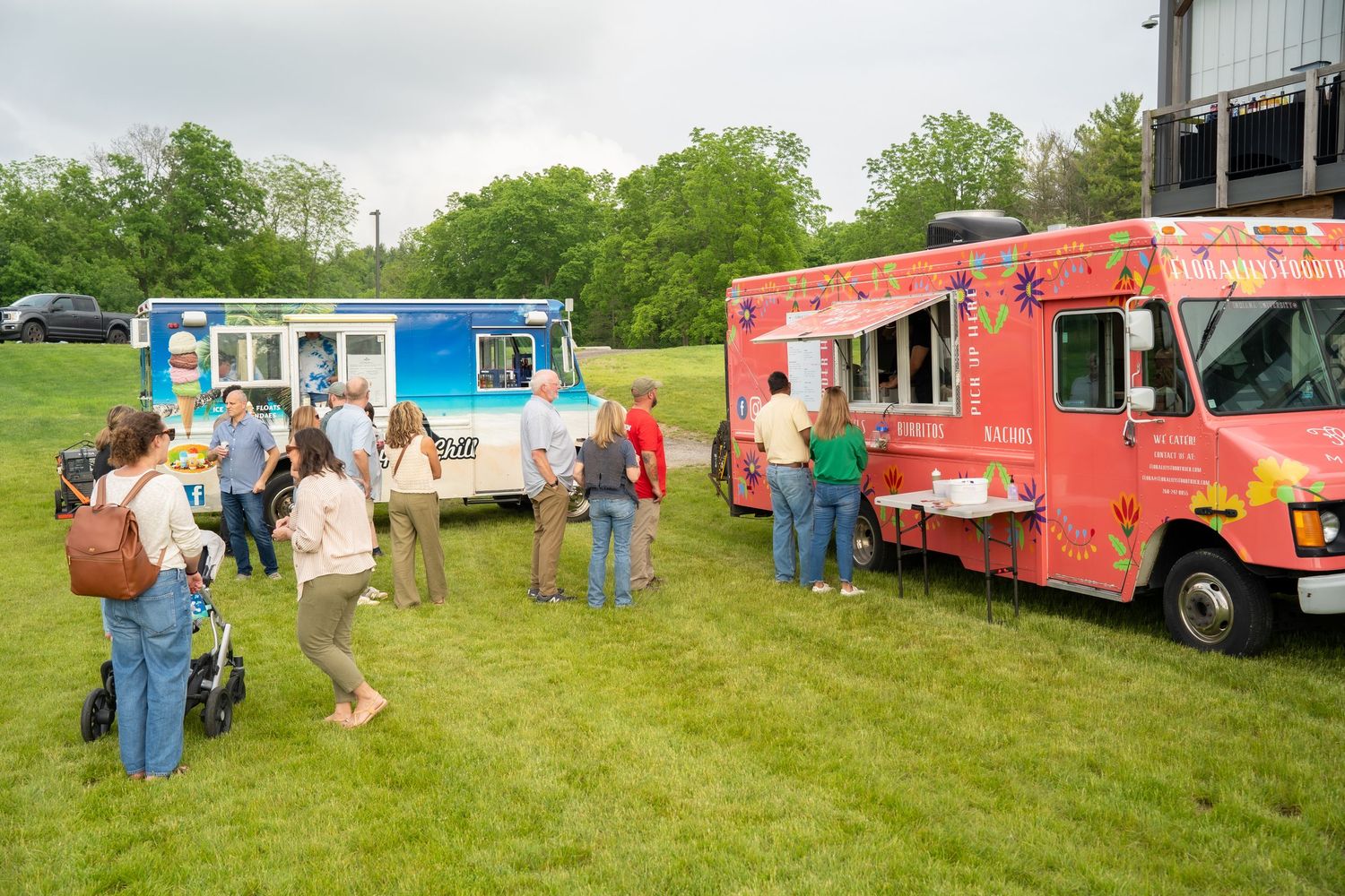 People lining up at two colorful food trucks parked on a green lawn at an outdoor community festival event.