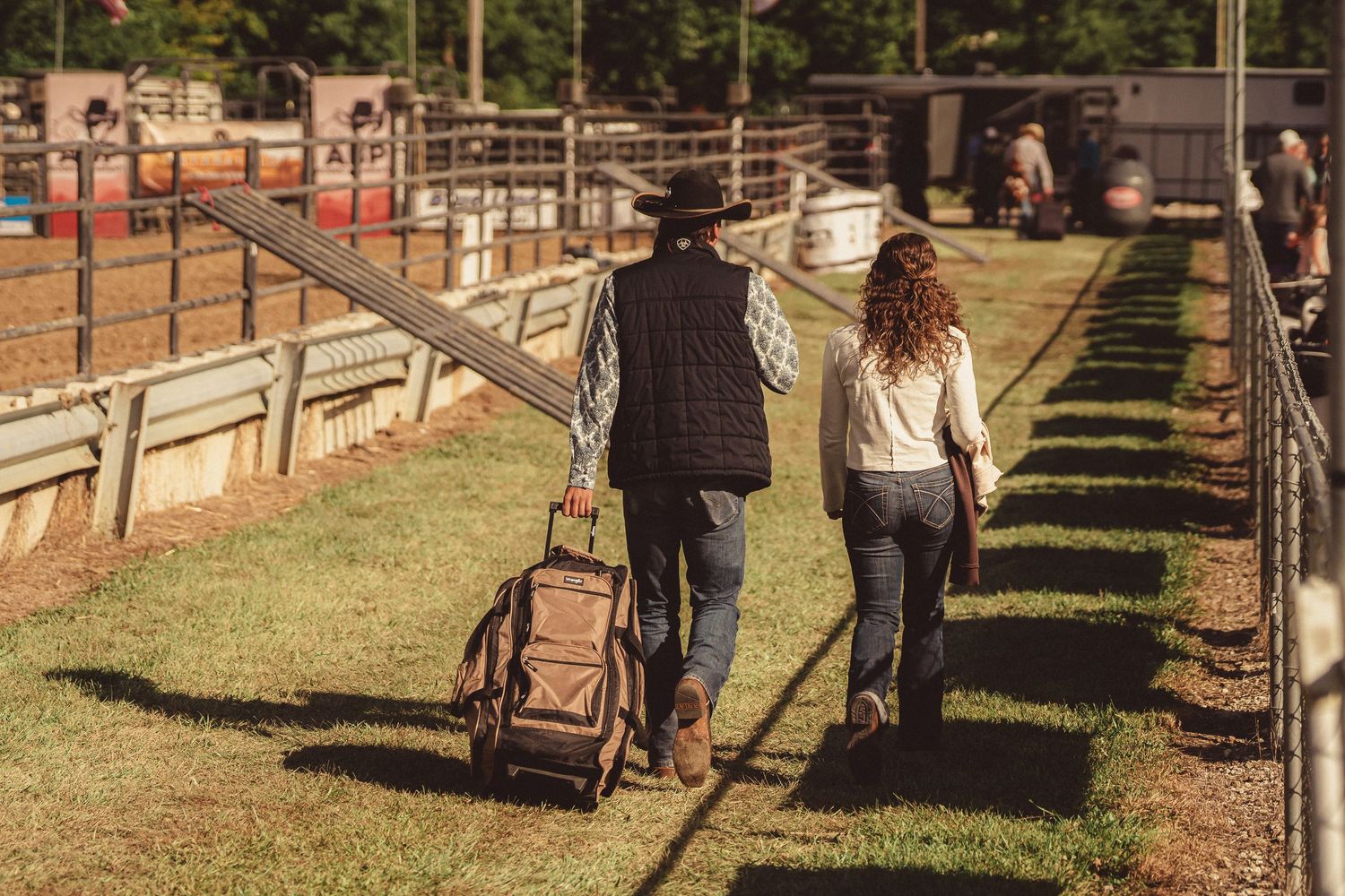 Cowboy and woman walking with luggage along fenced path at outdoor rodeo grounds.