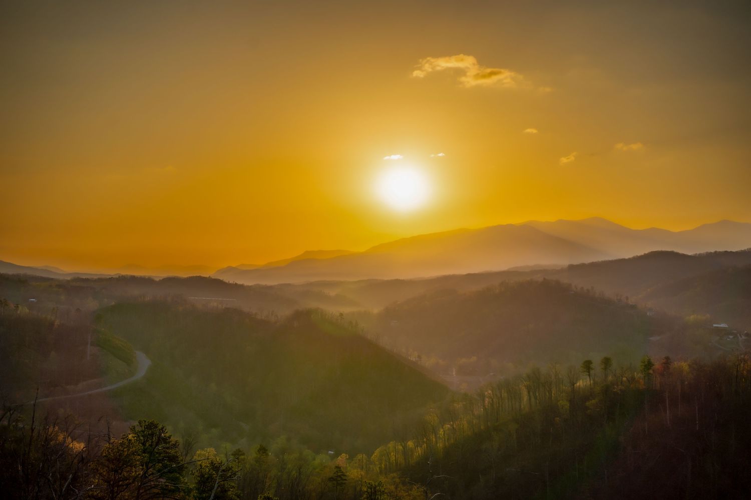 Golden sunset casts warm light over misty mountain valleys and winding road.