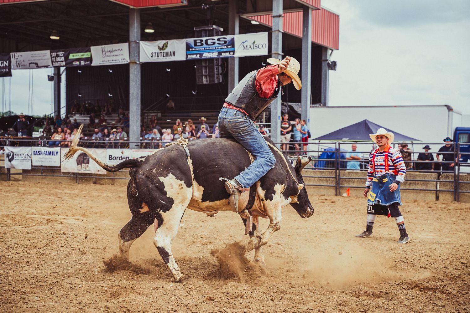 A cowboy rides a bucking bull at a rodeo arena while a clown stands nearby watching.