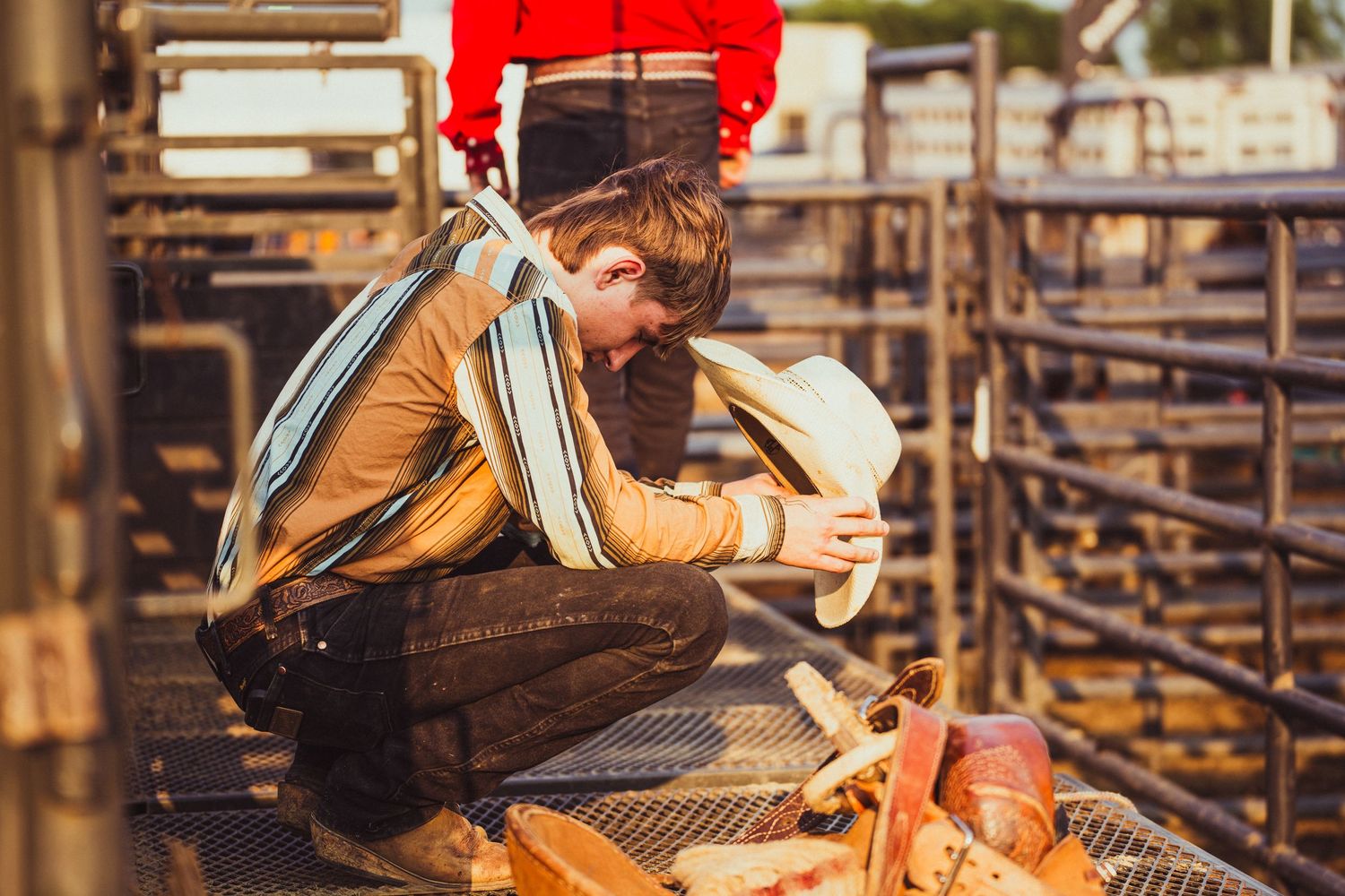 A cowboy crouches in a rodeo pen, examining his hat near equipment and metal fencing.